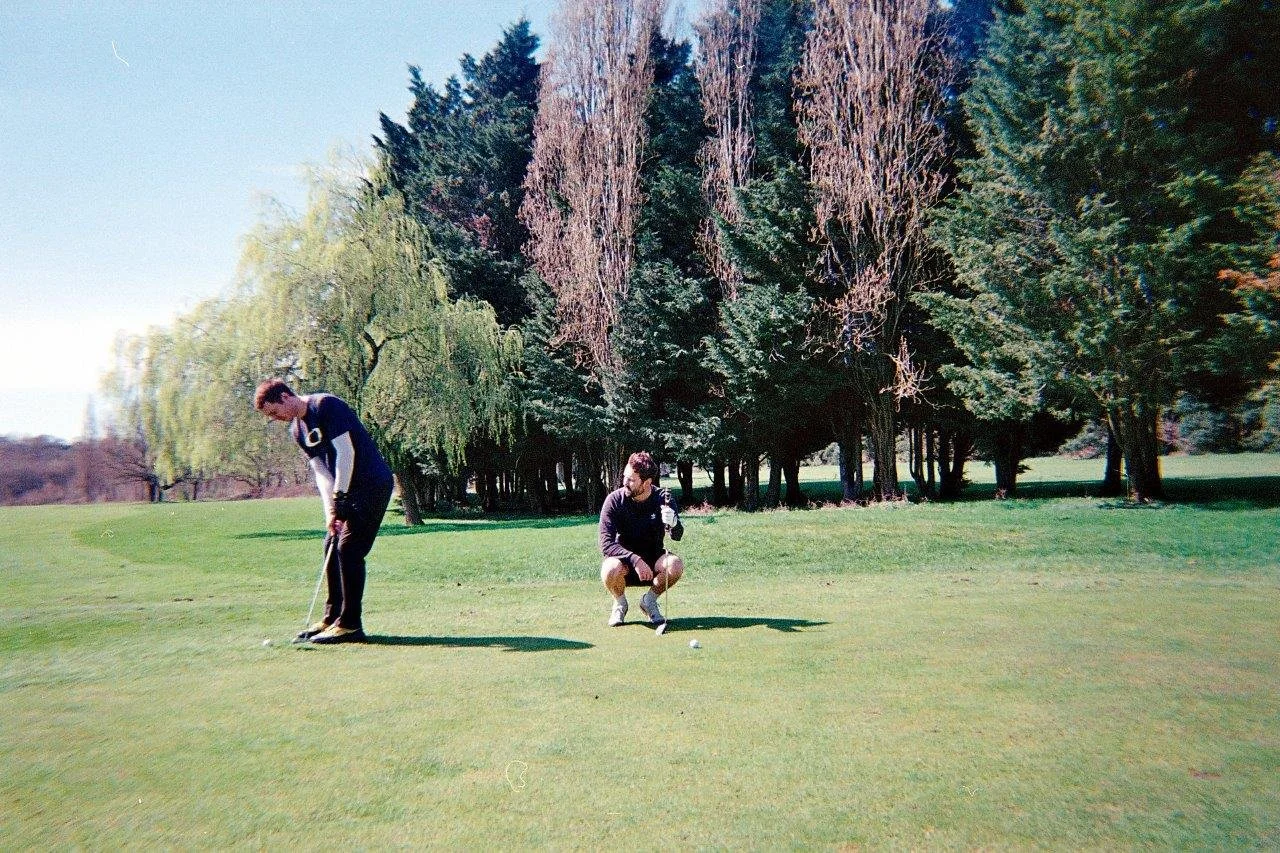 Two golfers on the putting green, one is about to putt, the other is squatting next to him watching.
