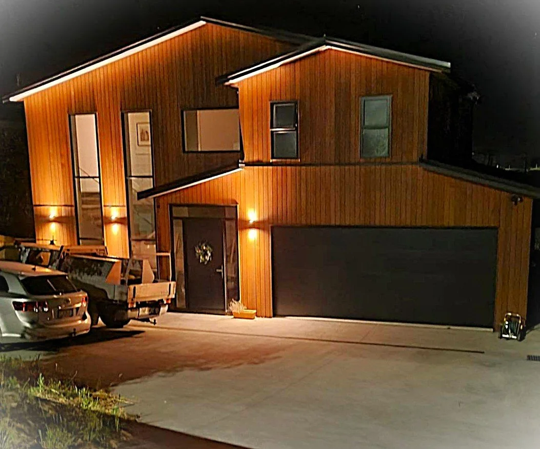 A two-story modern house at night with warm exterior lighting, wooden siding, large windows, and a black garage door. Three cars are parked in the driveway, and a small wreath decorates the front door.