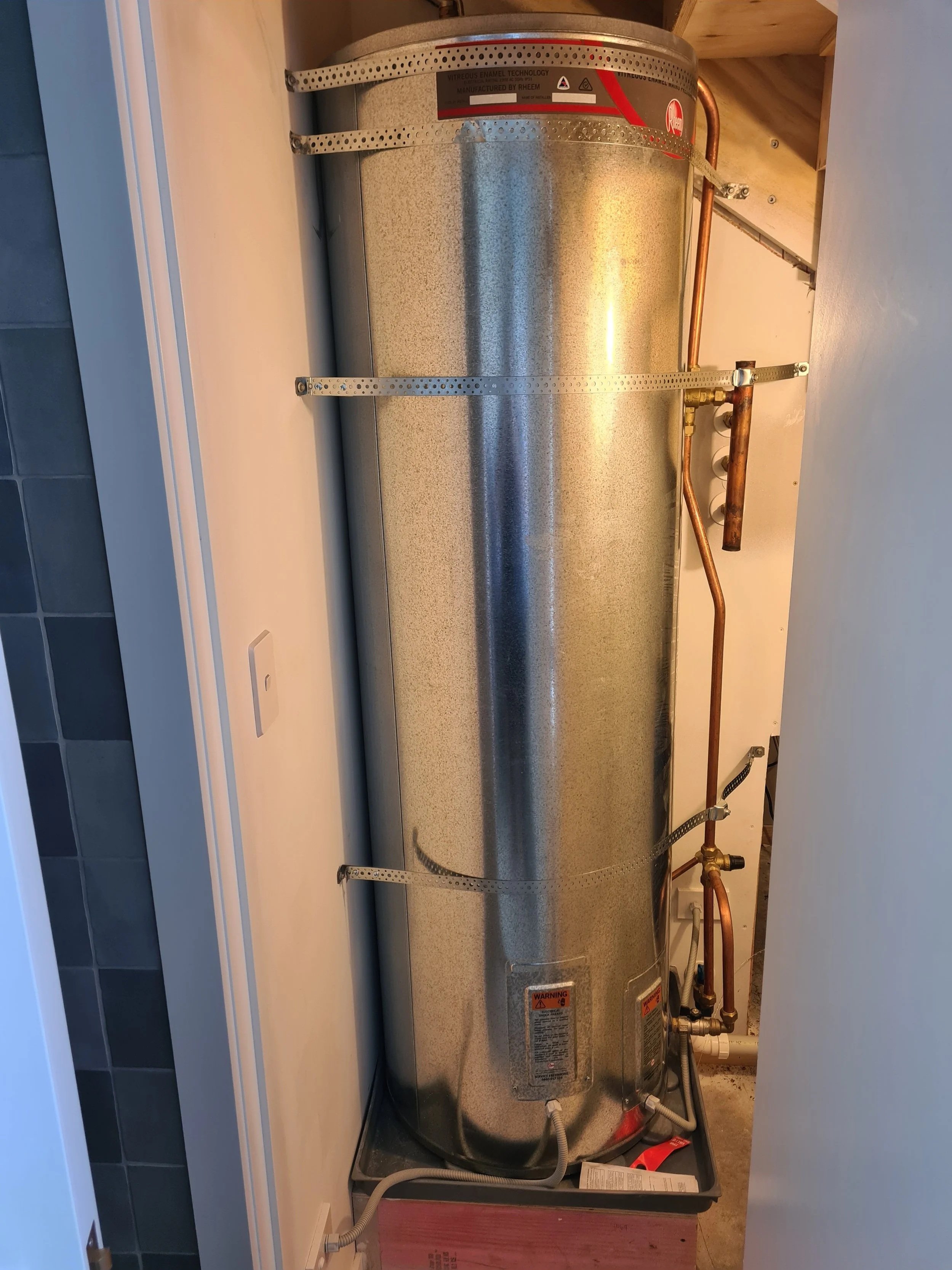 A large cylindrical water heater installed in a utility room with copper piping, mounted on the wall with metal straps.