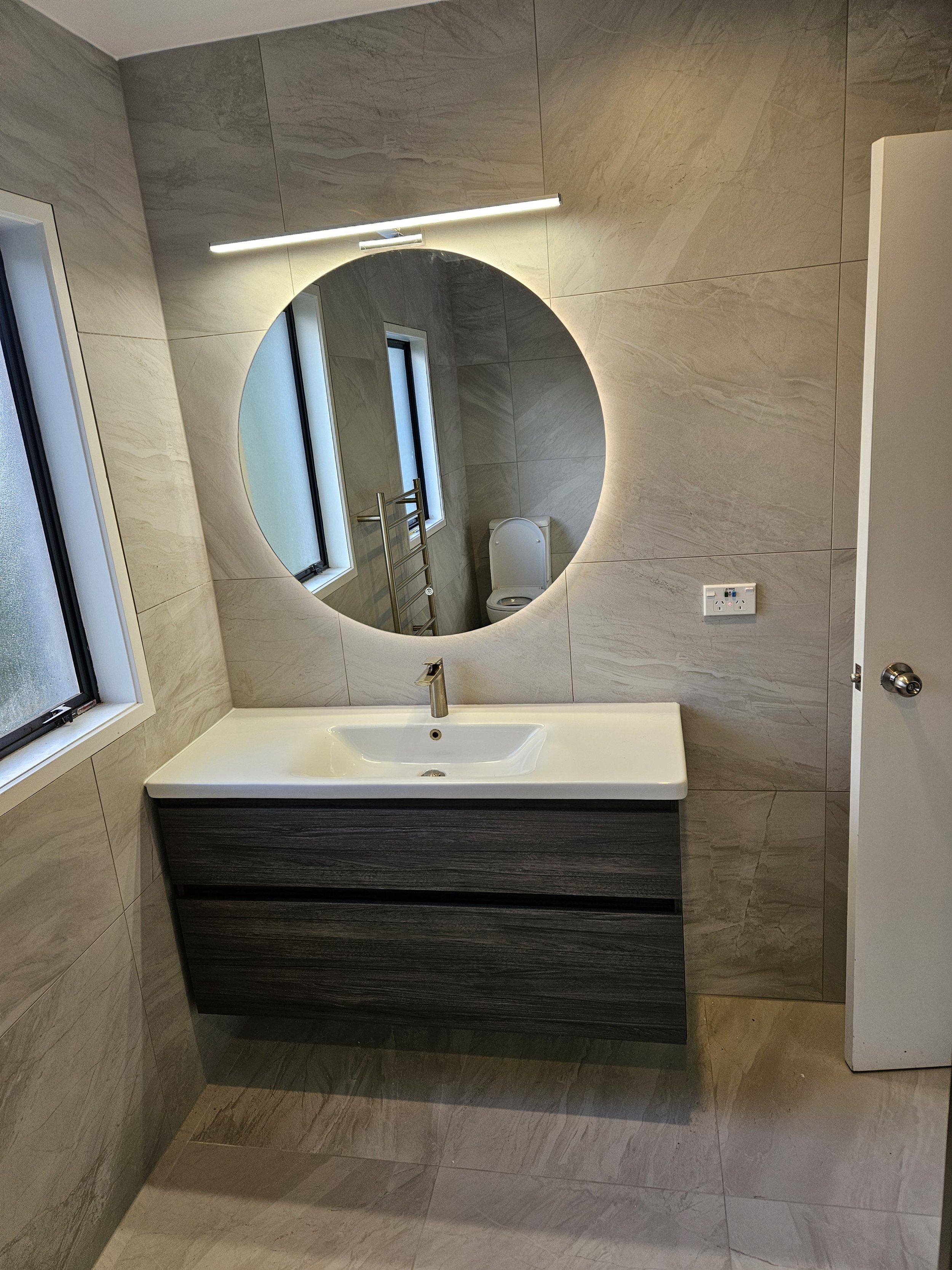Modern bathroom with a large circular backlit mirror above a white sink with a dark wood vanity, beige tiled walls, two narrow windows, a towel rack, and a toilet reflected in the mirror.