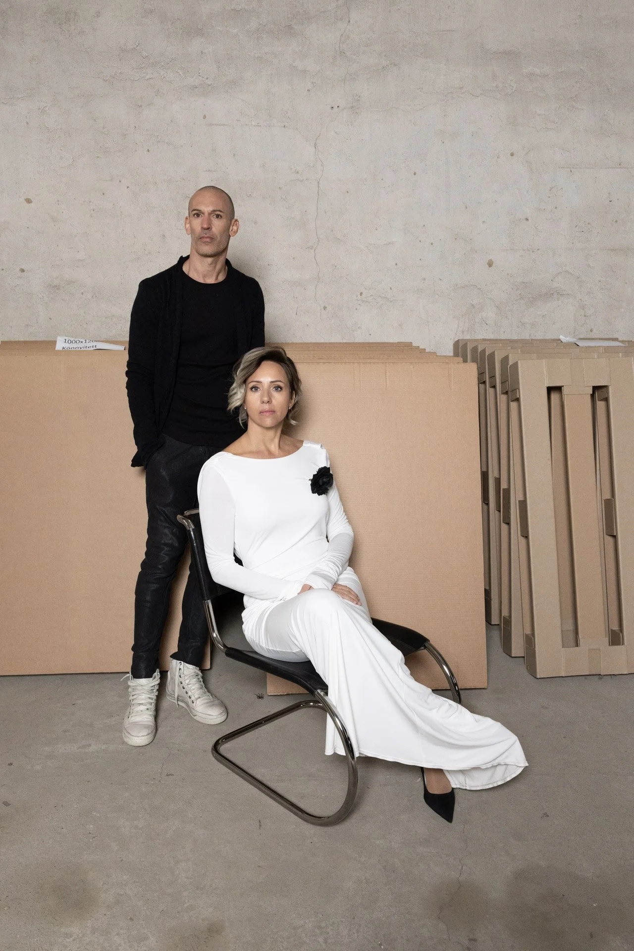 Two people indoors with cardboard boxes; one seated in a white dress and the other standing in black clothing next to them.