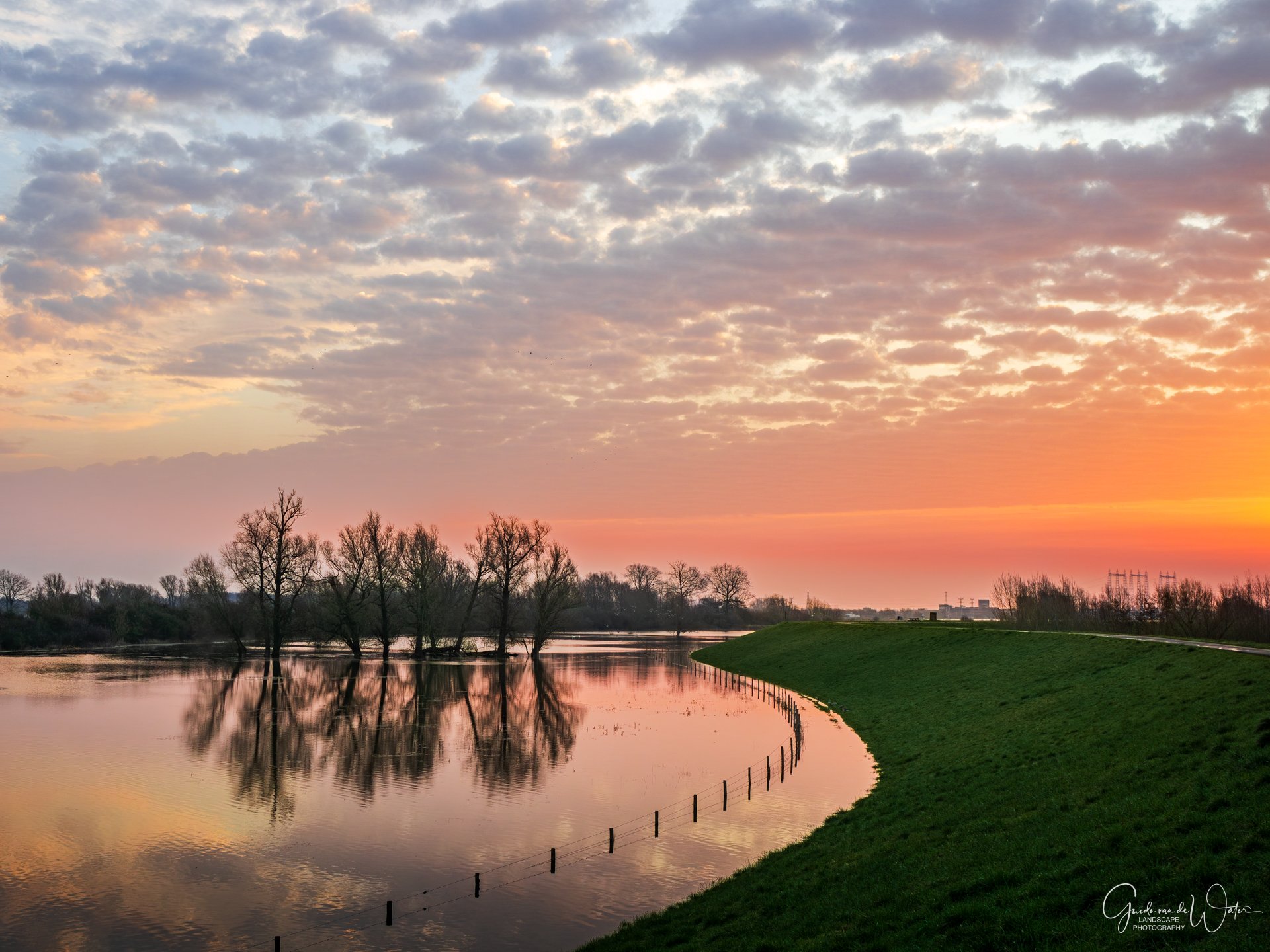Sunset over a river with trees on the bank and a grassy embankment, with a partly cloudy sky.