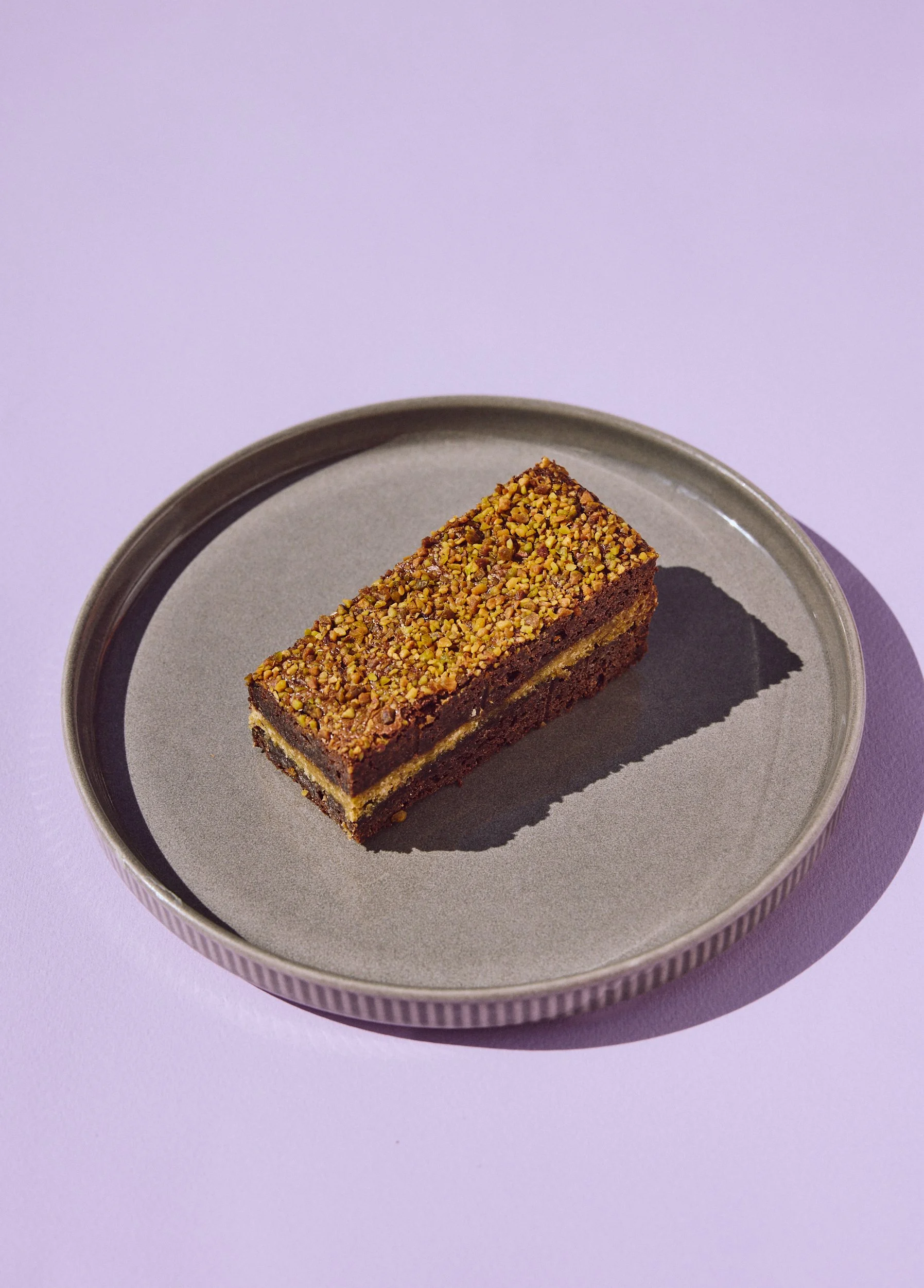 A rectangular layered dessert with a crumbly base, topped with chopped nuts, served on a round ceramic plate with a subtle striped rim, set against a pastel purple background.
