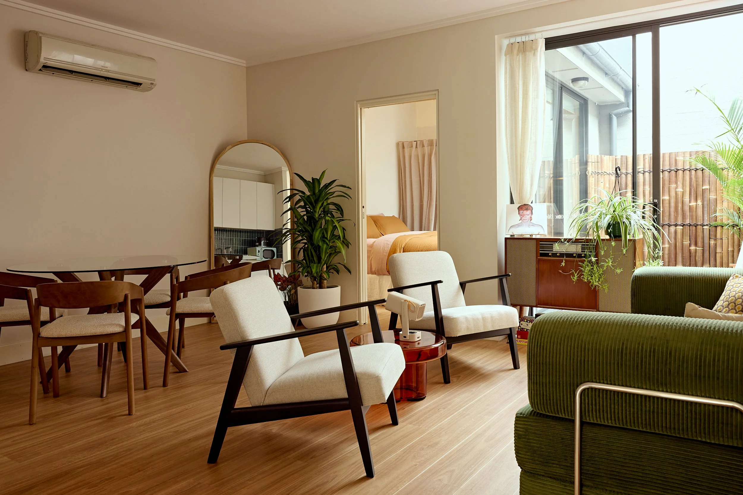 Living room with dining table, various chairs, a green armchair, potted plants near a large window with curtains, a mirror, and a glimpse of a bedroom with a bed and curtains.