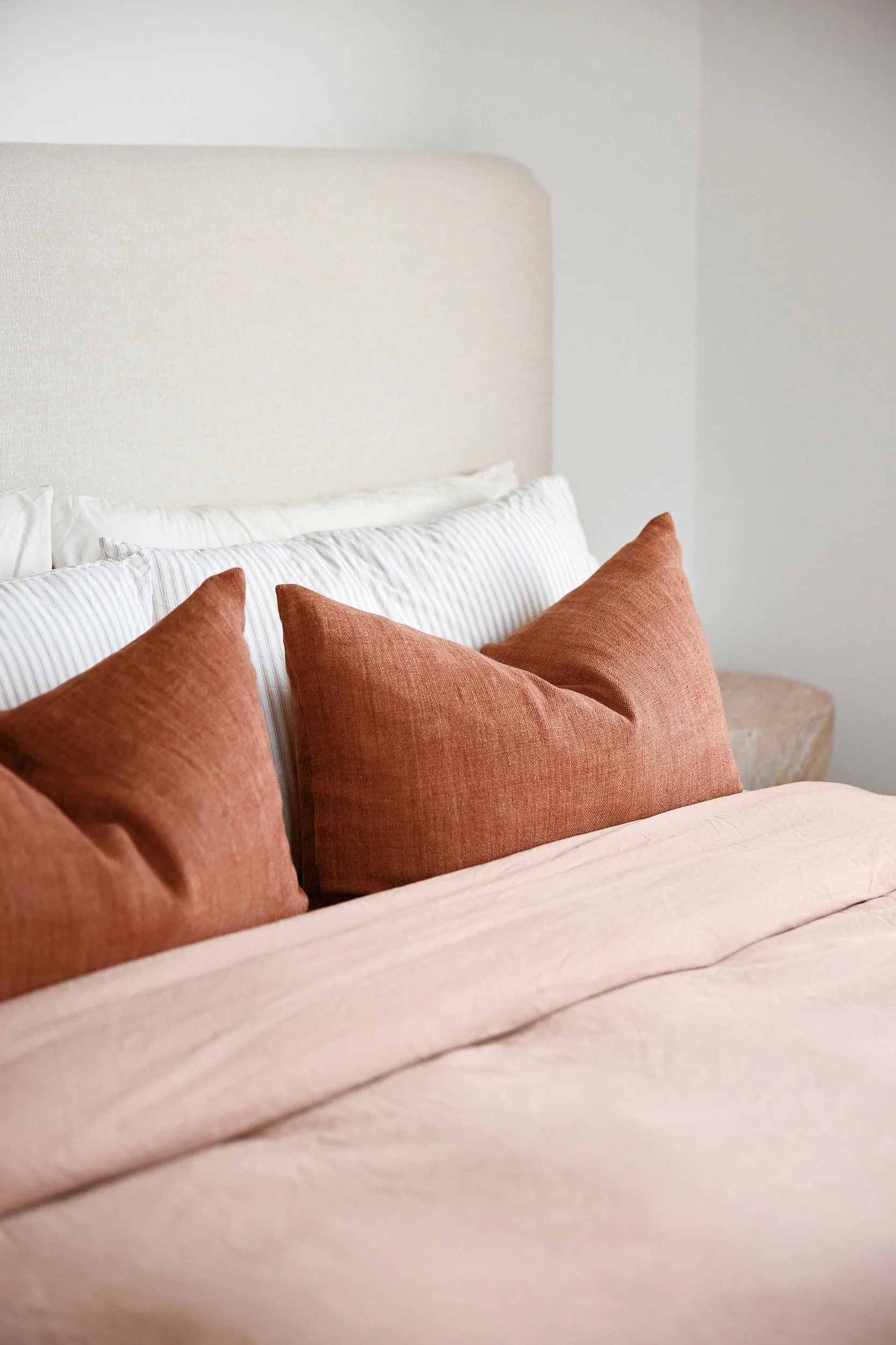 A close-up of a cozy bedroom with a bed featuring a pink duvet and rust-colored pillows. The background shows a white headboard and white bedding, with a neutral wall and a small wooden side table.