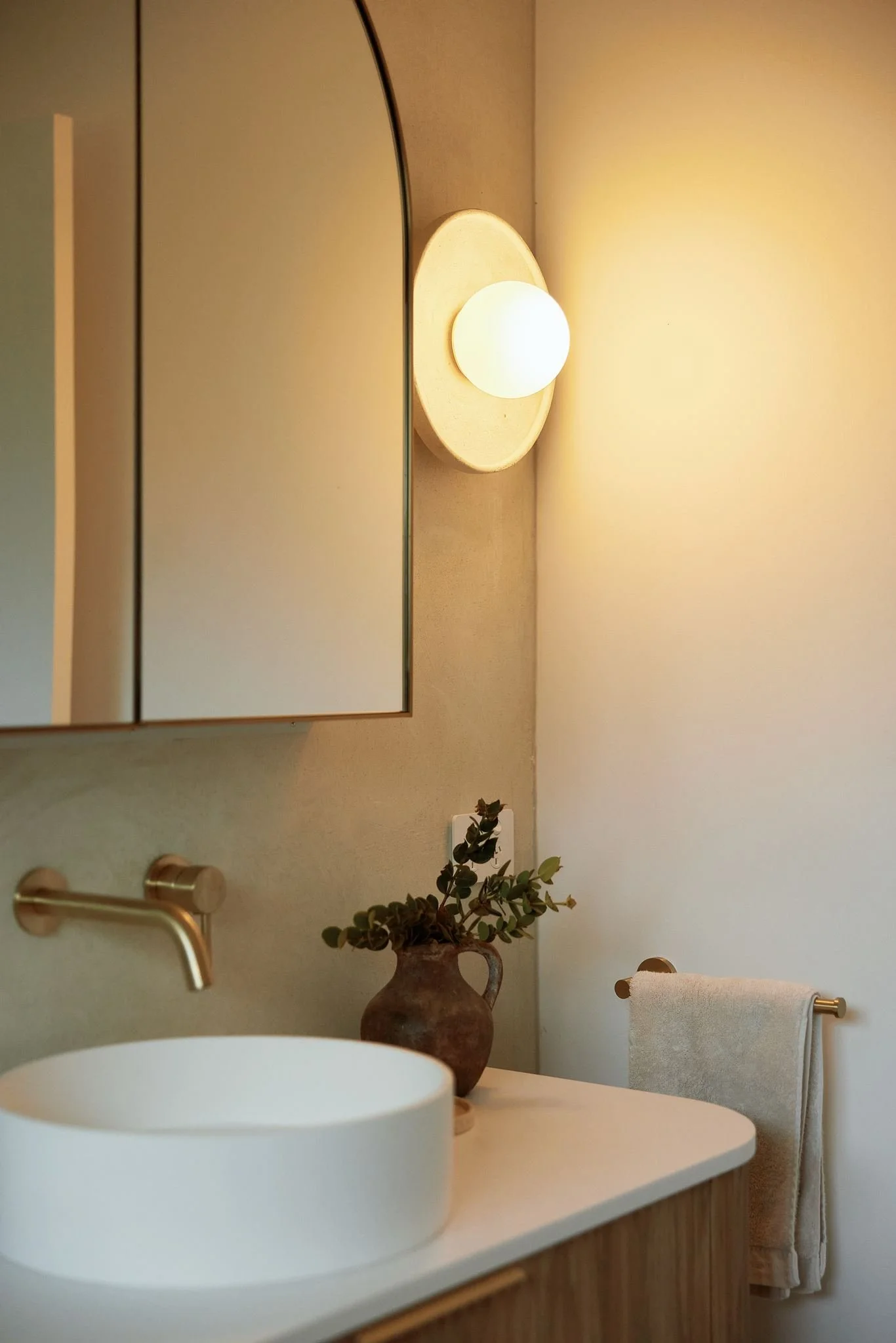 A modern bathroom with a round white vessel sink on a wooden vanity, a brass faucet, a mirror, a wall-mounted light, a decorative vase with green foliage, and towel racks with a white towel.