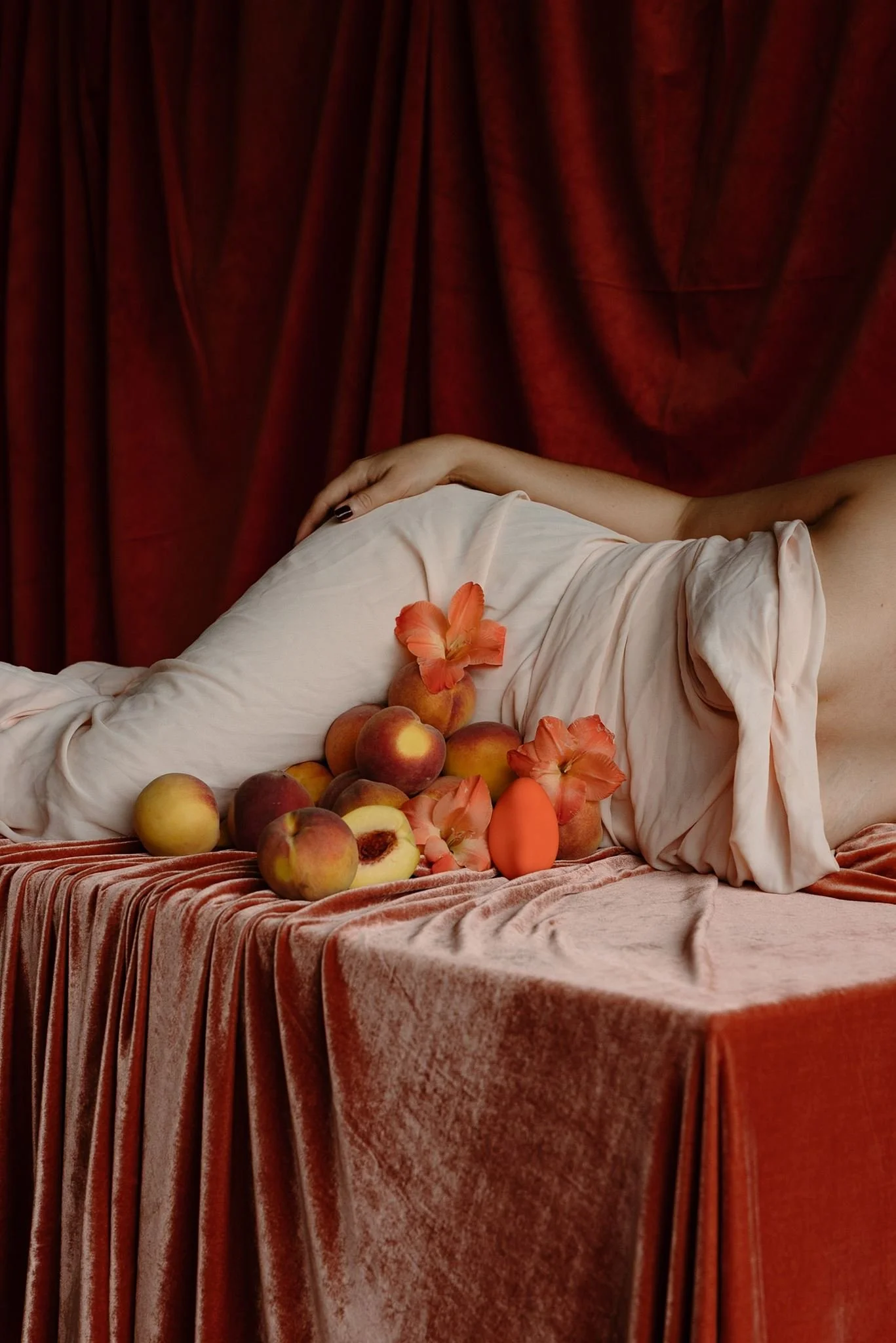 A female lying on a bed with a beige blanket, surrounded by peaches and peach blossoms, with a deep red curtain in the background.