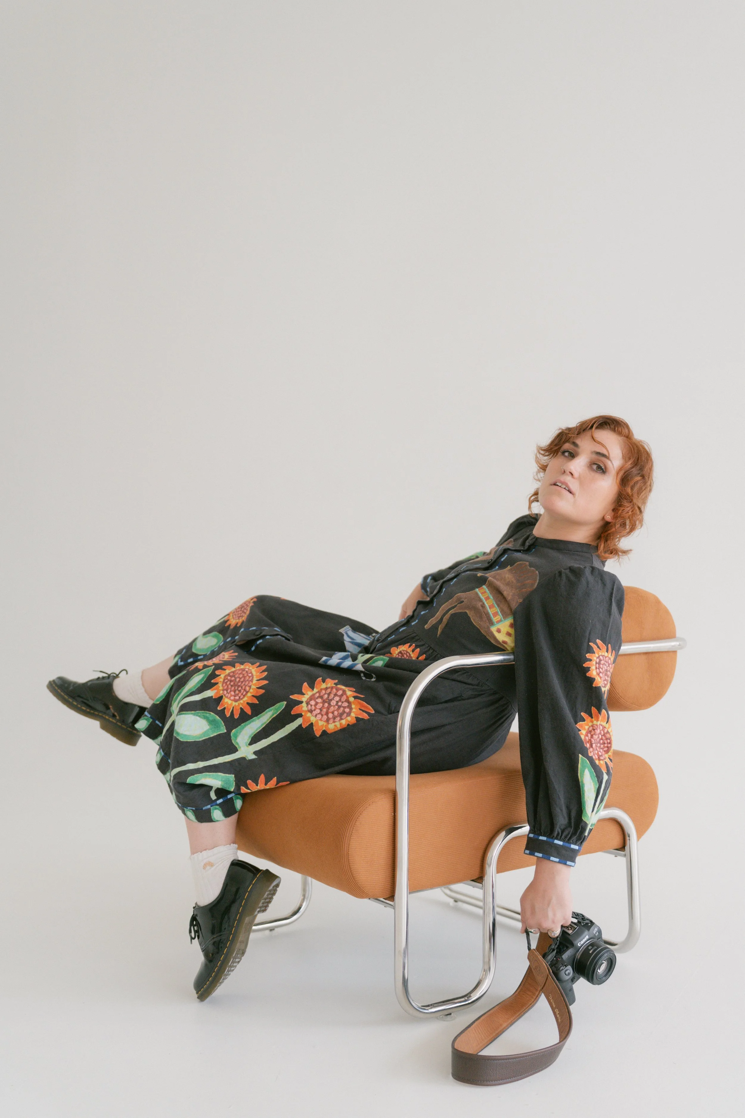 Young woman with short curly red hair sitting on a vintage orange chair, wearing a black dress with a colorful sunflower pattern, holding a camera, and looking at the camera with a relaxed expression against a plain white background.