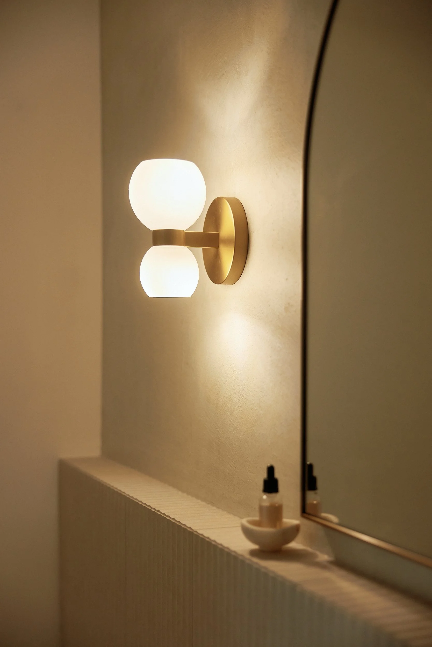 Close-up of a modern wall light fixture with two frosted glass globes mounted on a gold-colored base, illuminating a beige wall in a room with a mirror and small bottles on the countertop.