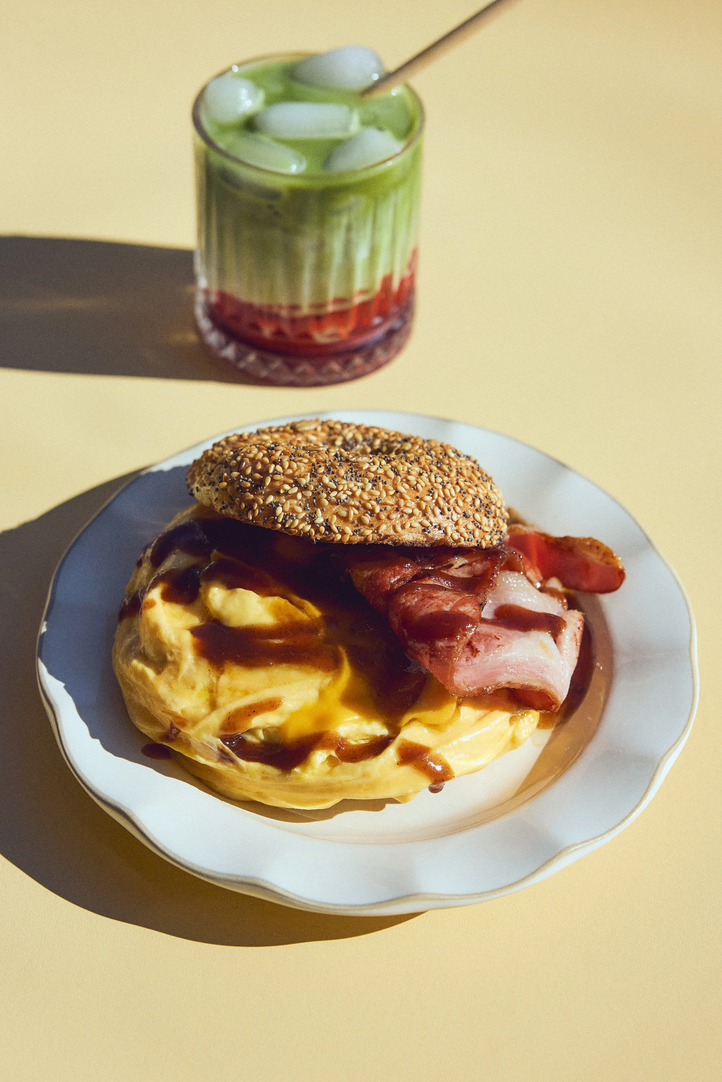 Close-up of a plate with a breakfast scramble topped with bacon and gravy, and a glass of green iced drink with ice cubes and a straw on a yellow background.