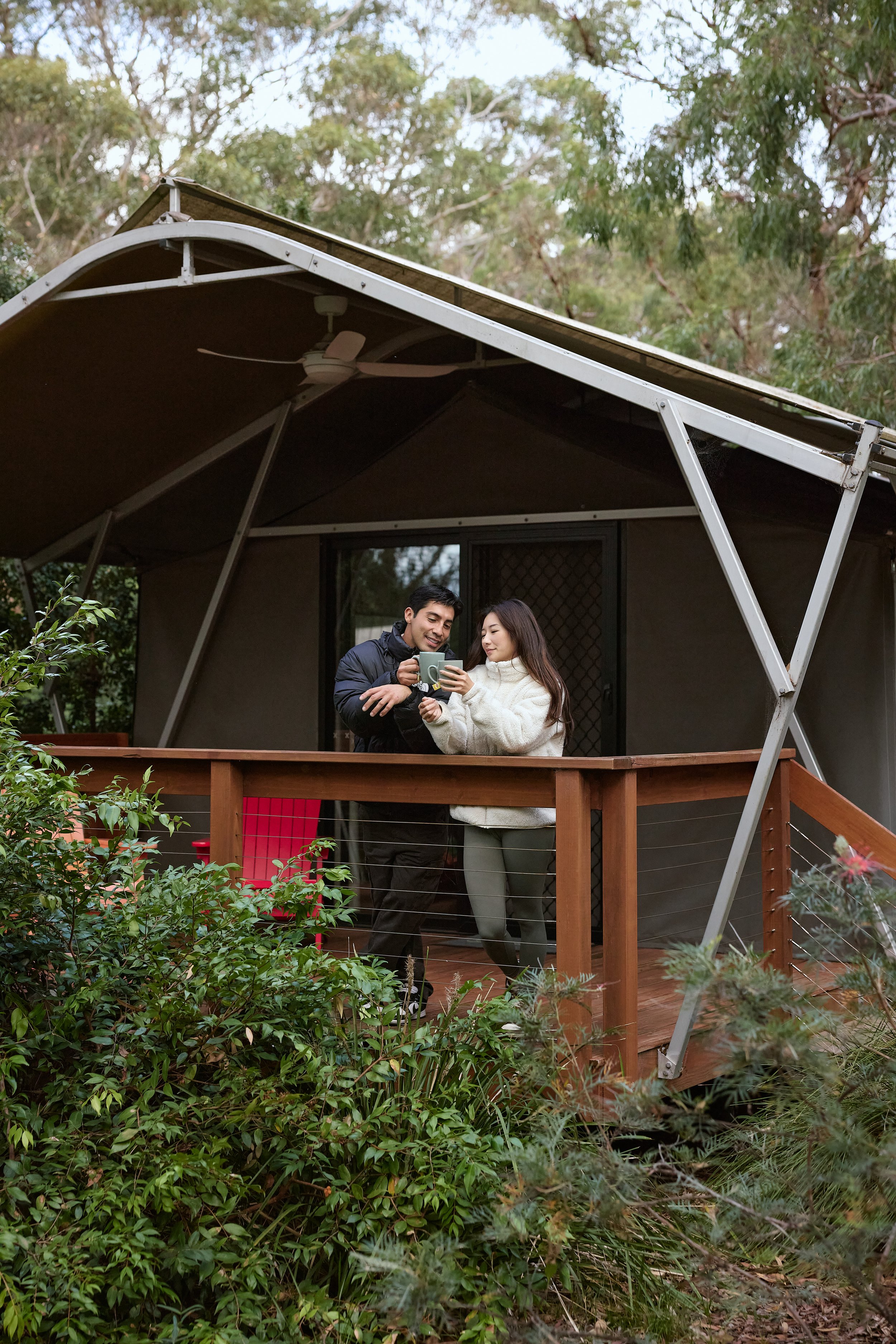 A man and woman on a wooden porch of a tiny house, surrounded by greenery, looking at something on a smartphone together