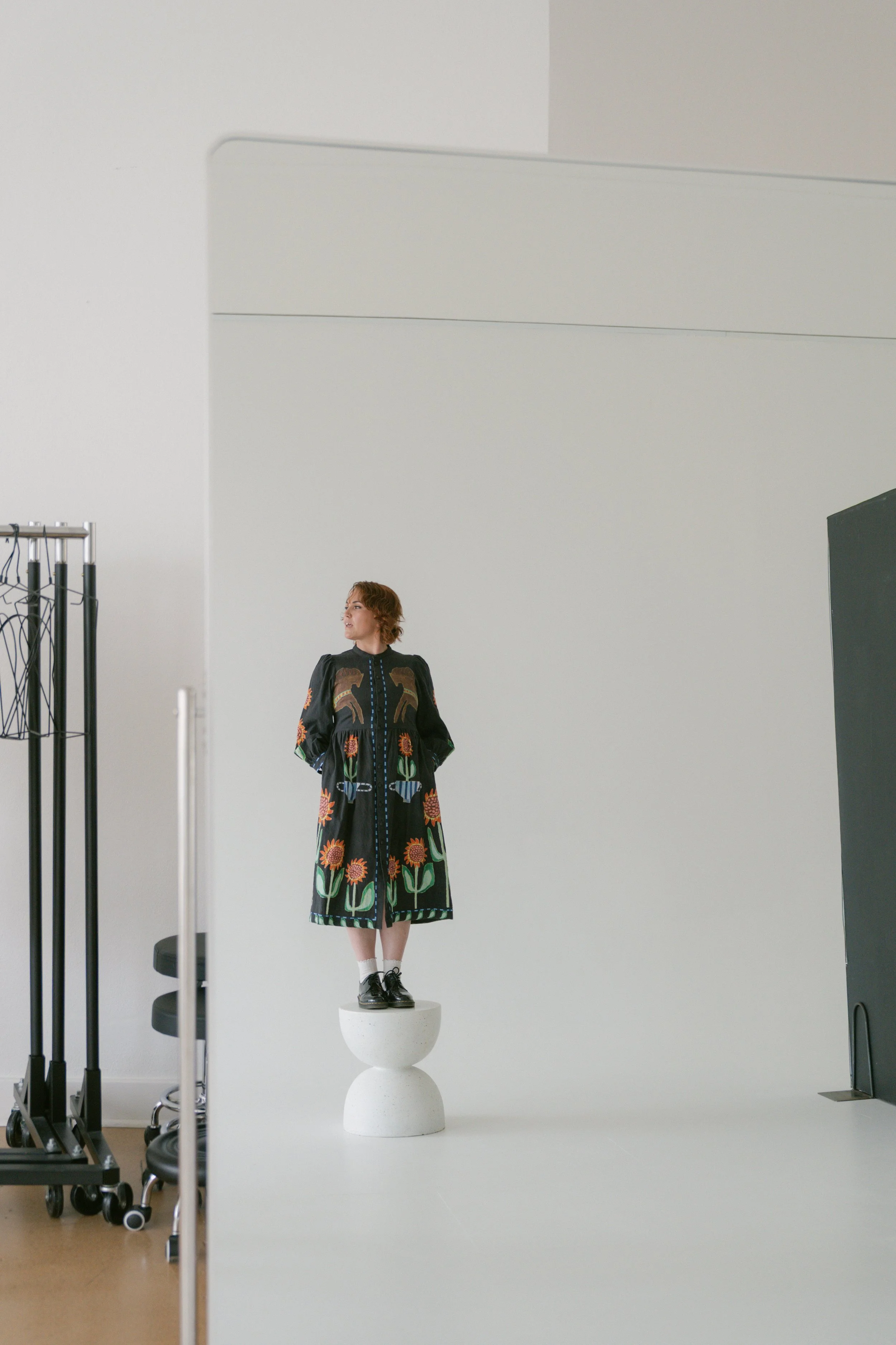 A woman with short red hair wearing a black embroidered dress with floral and animal designs, standing on a white pedestal in a photography studio with minimalistic decor.