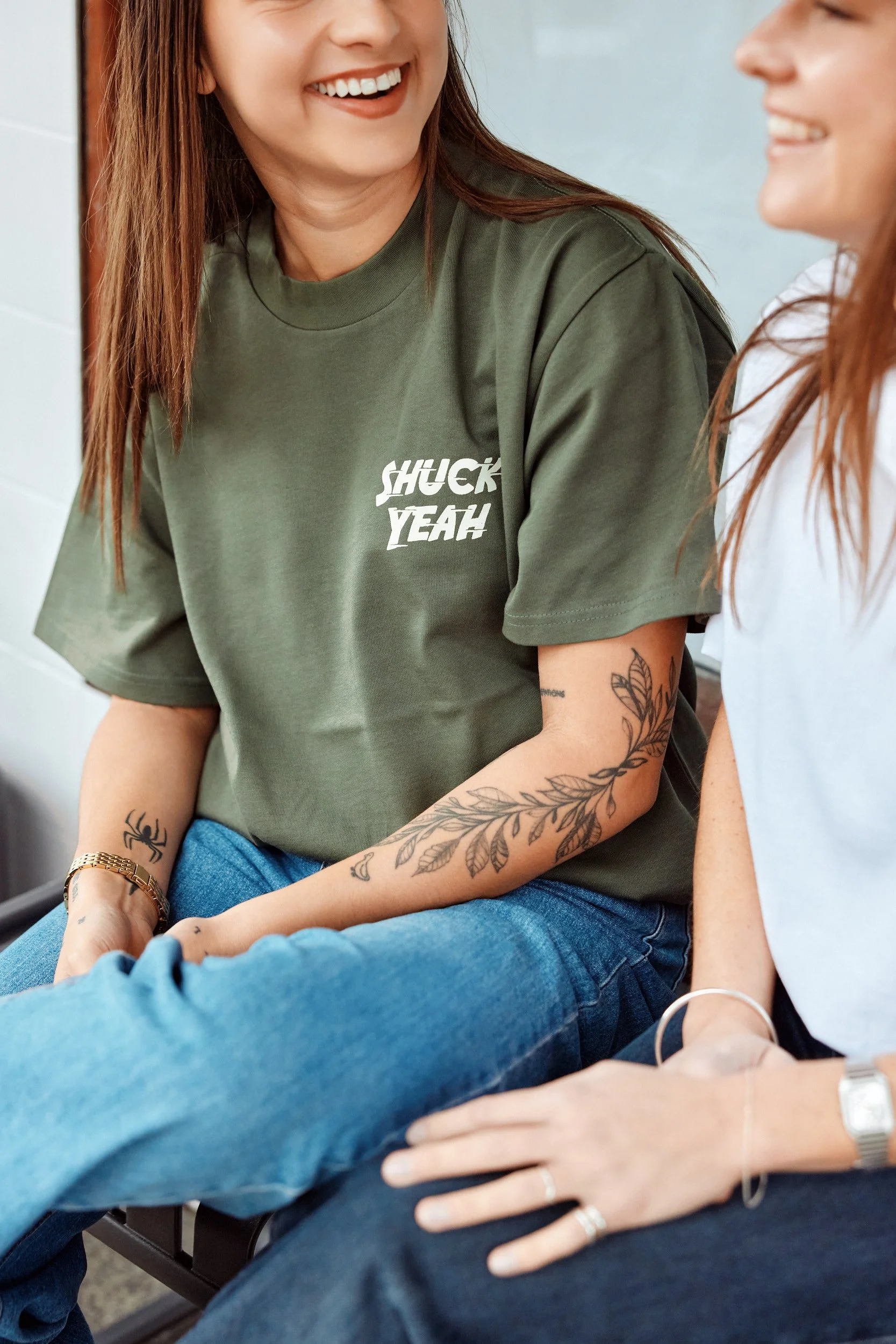 Two women sitting together, smiling, with tattoos on their arms, wearing casual clothes. One in an olive green shirt with the words 'SHUCK YEAH!' and the other in a white shirt. They are sitting close, likely in a casual or social setting.