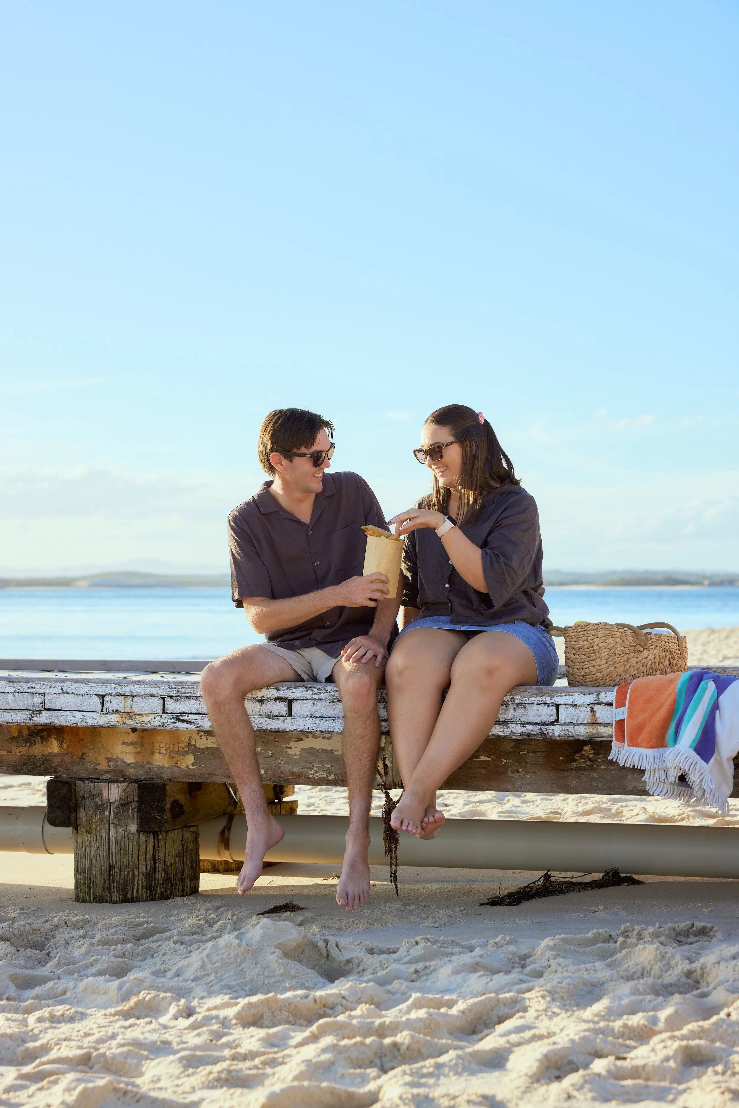 A young man and woman sitting on a wooden dock by the beach, smiling and sharing a snack together, with a blue sky and water in the background.
