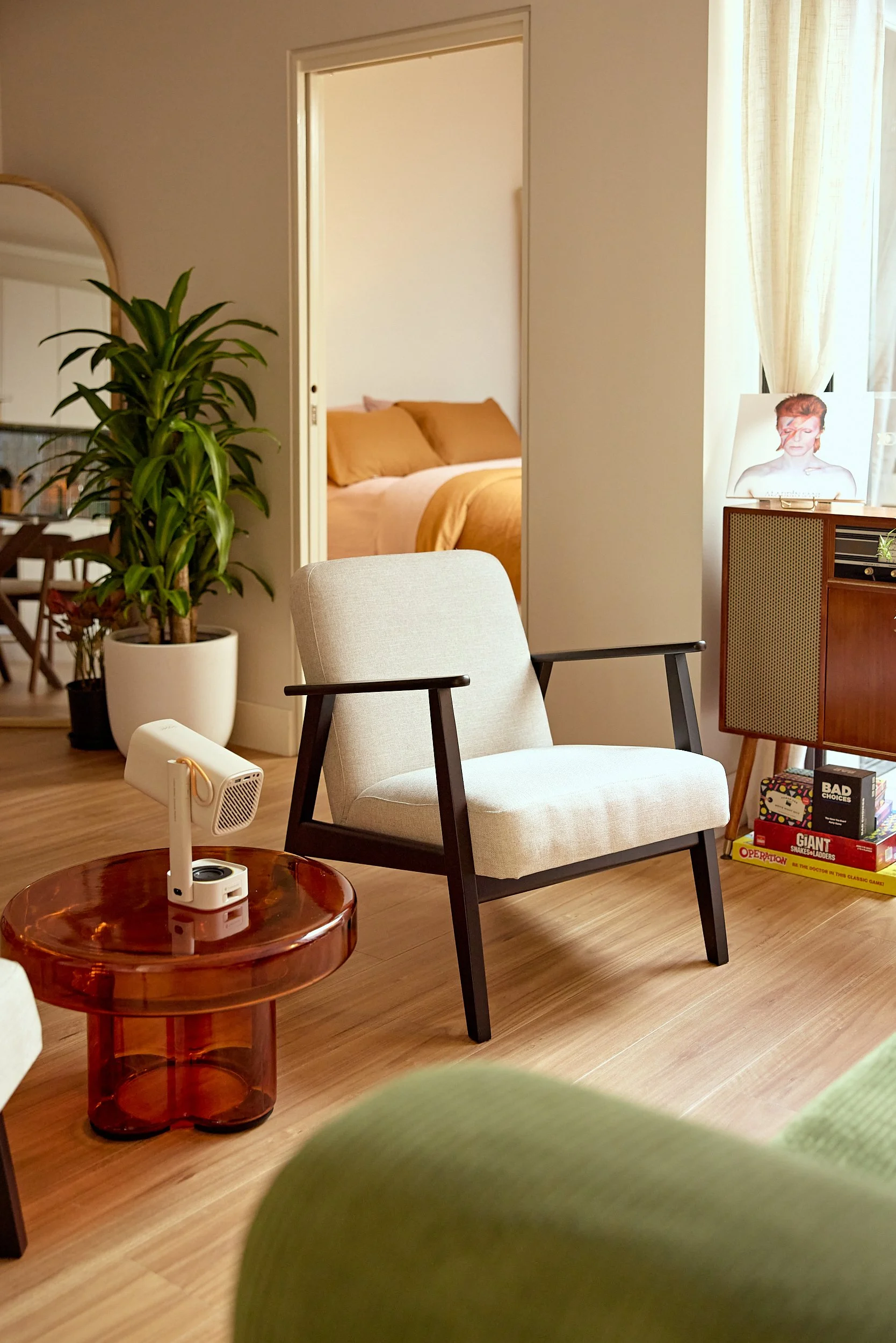 Living room with a white armchair, a small round wooden side table with a white device on it, a tall potted plant, a bookshelf filled with games, a doorway leading to a bedroom with a made bed, a framed photo of David Bowie, and a window with curtains.