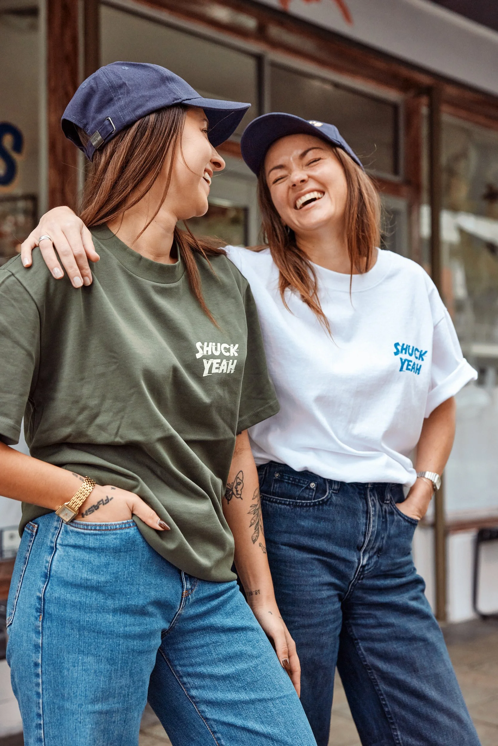 Two women standing outdoors, wearing casual t-shirts and baseball caps, smiling and talking to each other.