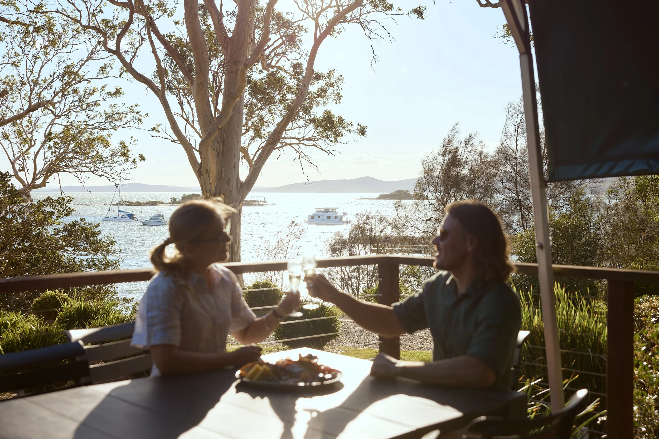 Two women sitting at an outdoor table, holding glasses in a toast near a waterfront with boats and trees in the background.