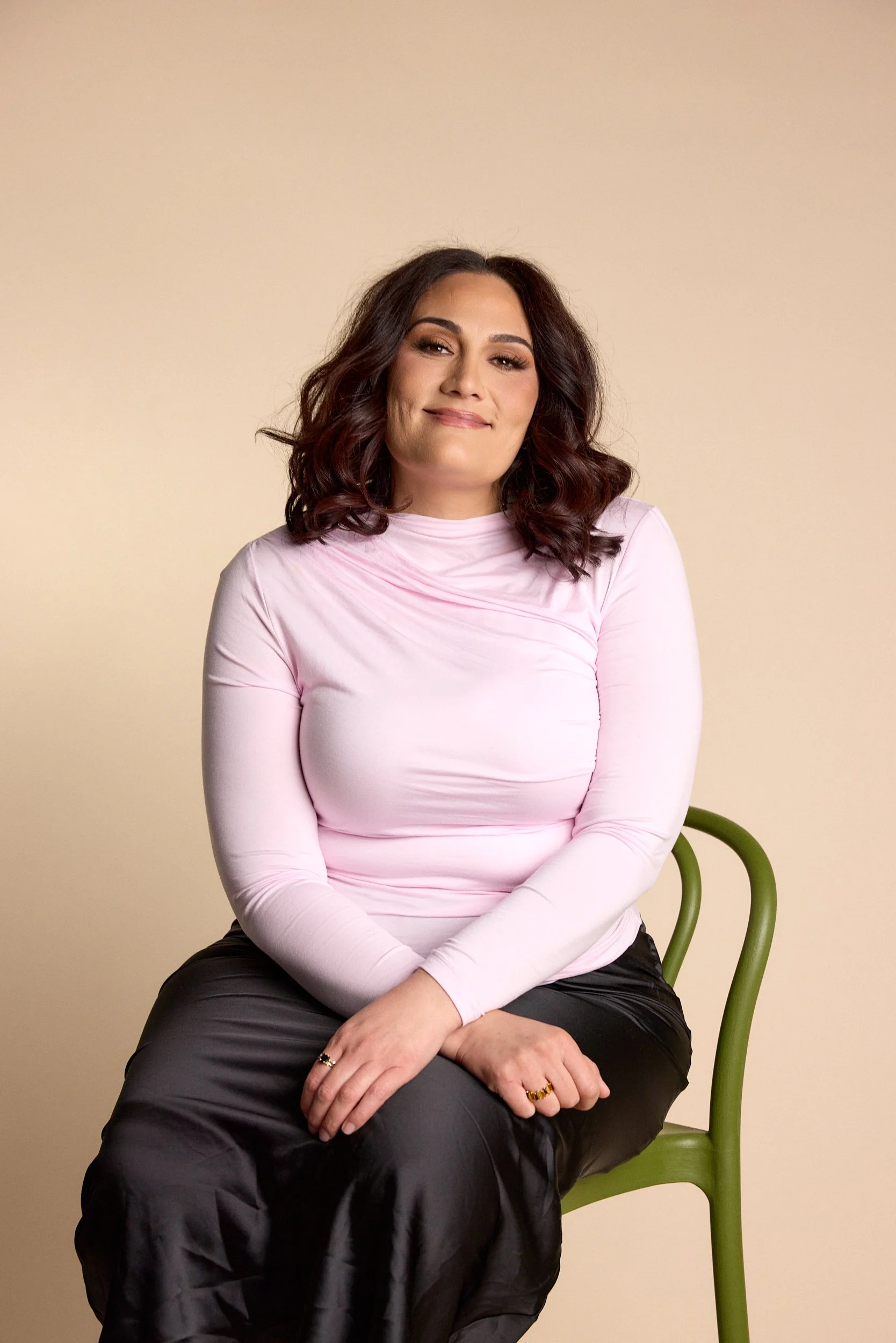 A woman with dark wavy hair sitting on a green chair in front of a plain beige background. She is wearing a light pink long-sleeve top and dark pants, smiling softly.