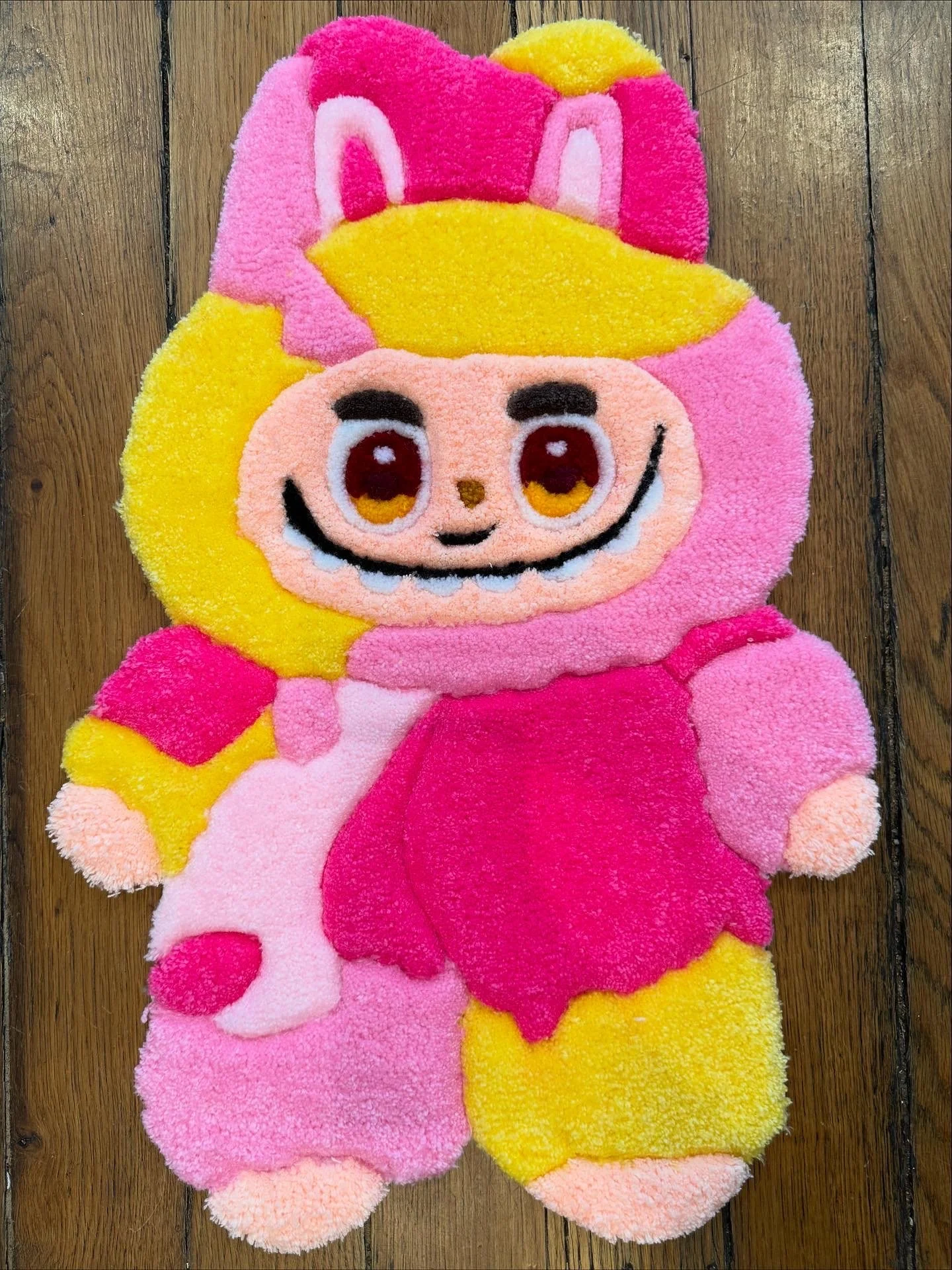 Tapis en forme de personnage de dessin animé avec un visage souriant, des grands yeux, des cheveux blonds et une robe rose, posé sur un sol en bois.