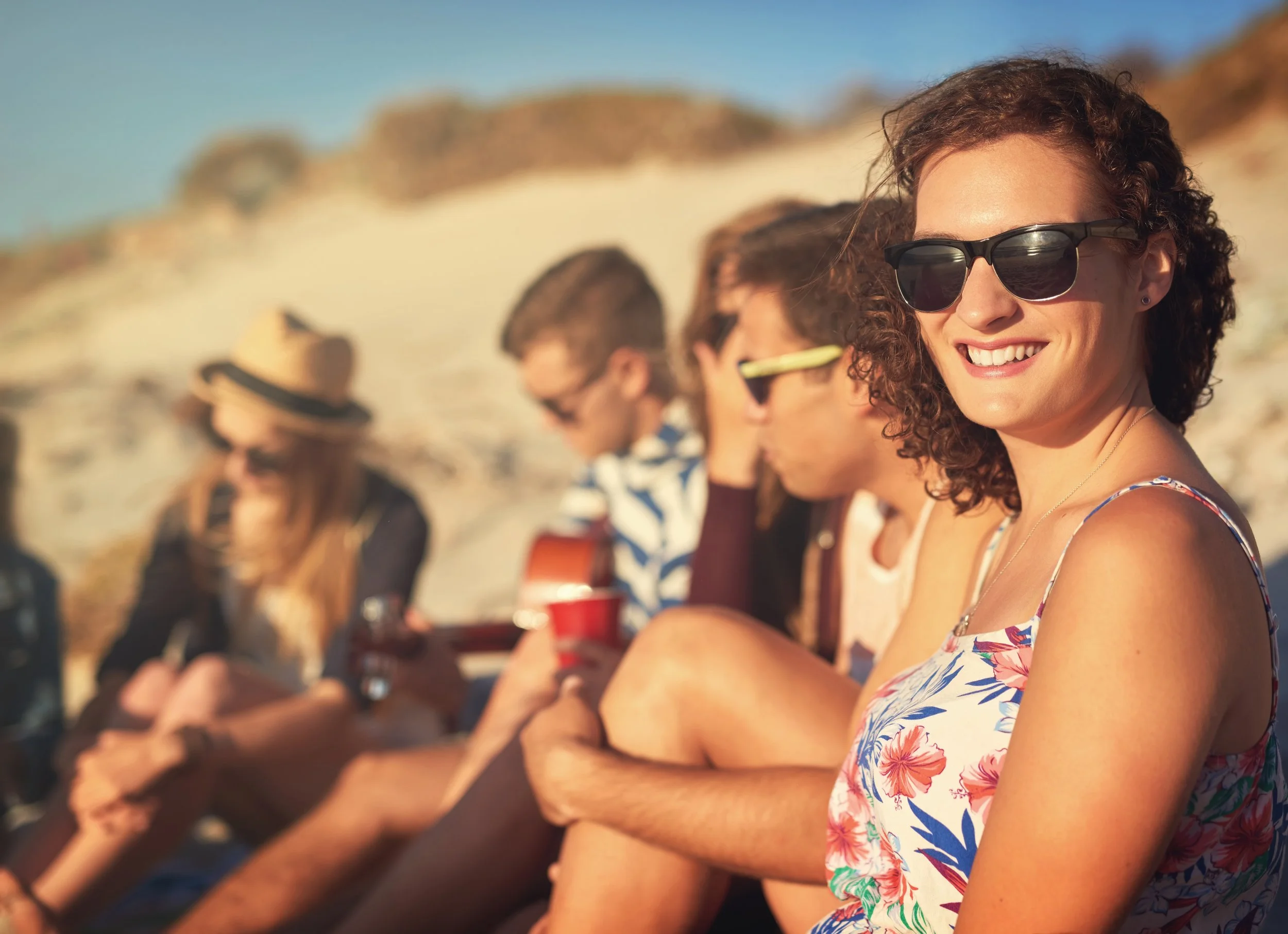 : Friends relaxing on a sunny beach, with a woman in sunglasses smiling in the foreground.