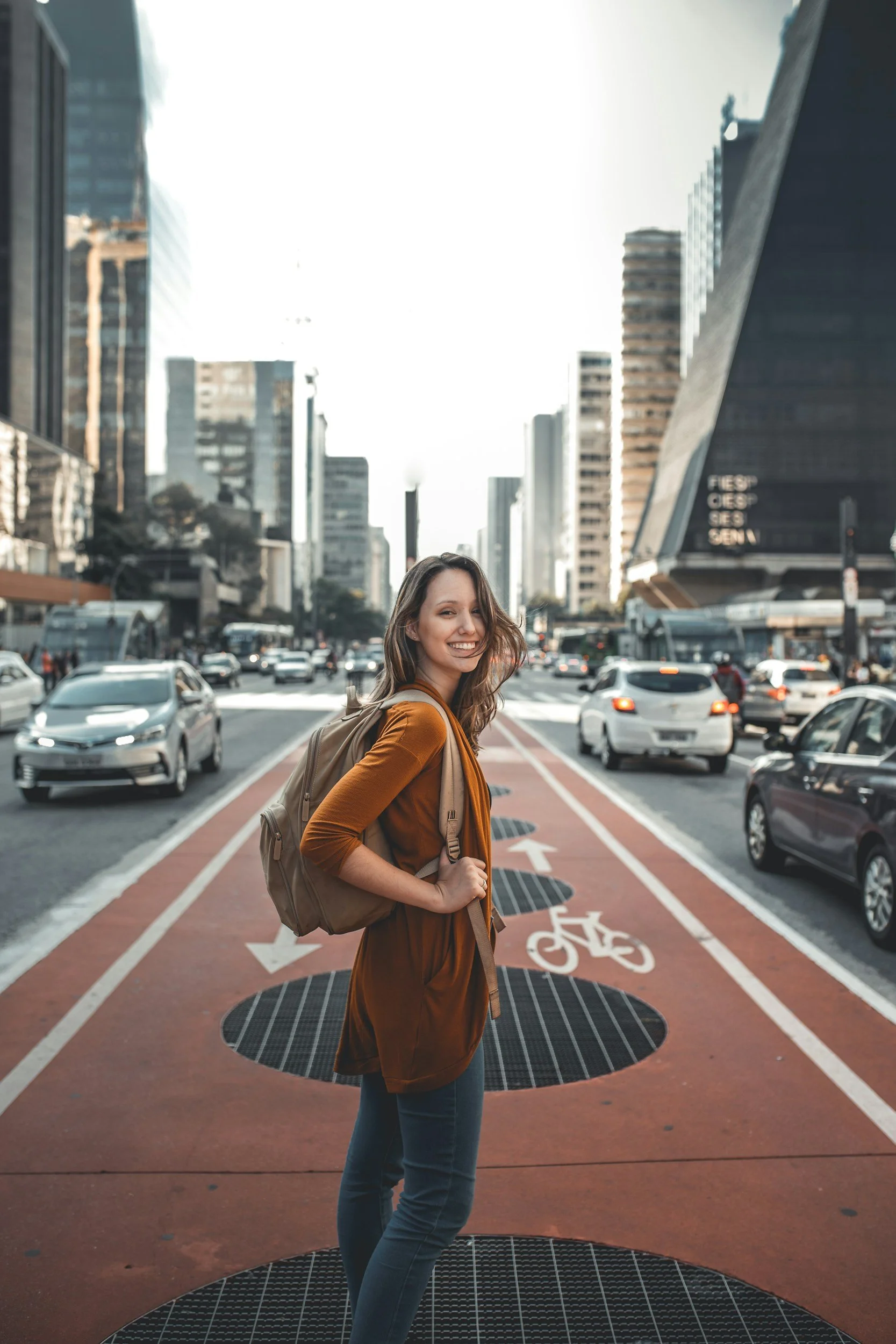 Young solo traveler with a backpack standing on a city bike lane, smiling at the camera with tall modern buildings and traffic in the background.