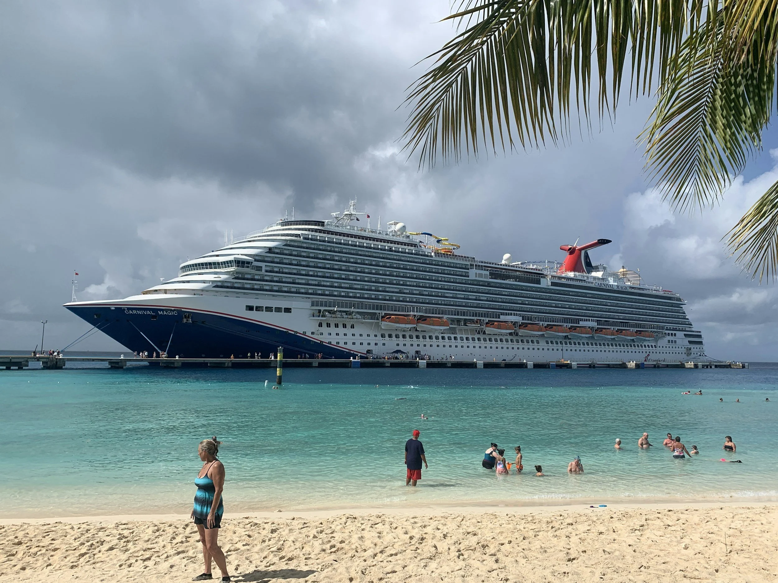 Woman standing on a sandy beach looking toward a large cruise ship anchored in turquoise water under a cloudy sky, with swimmers in the sea and palm leaves framing the scene.
