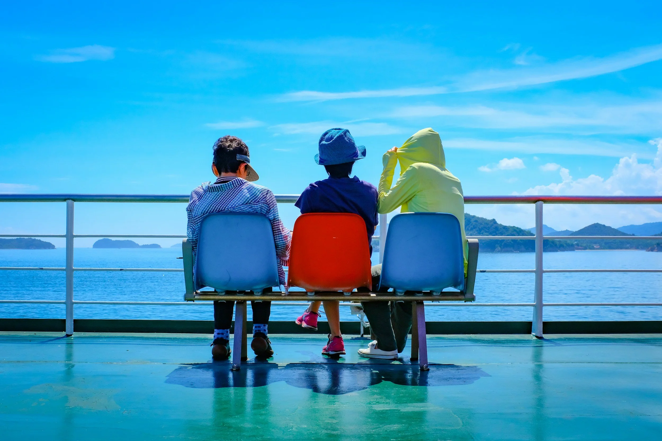 “Three kids sitting on colorful chairs on a ferry deck, looking out at the blue sea and distant islands on a sunny day.”