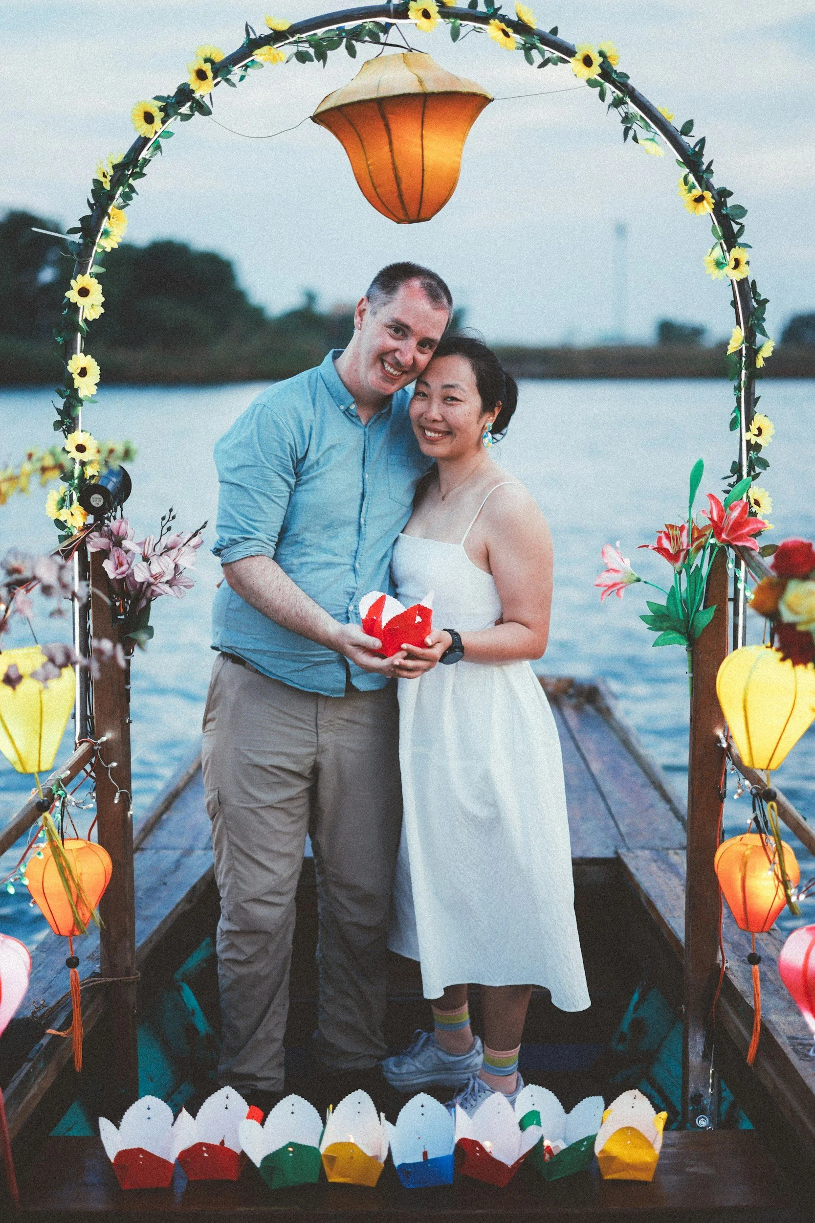 Smiling couple standing on a small decorated boat at sunset, surrounded by colorful lanterns and flowers on a calm river – romantic honeymoon-style scene.