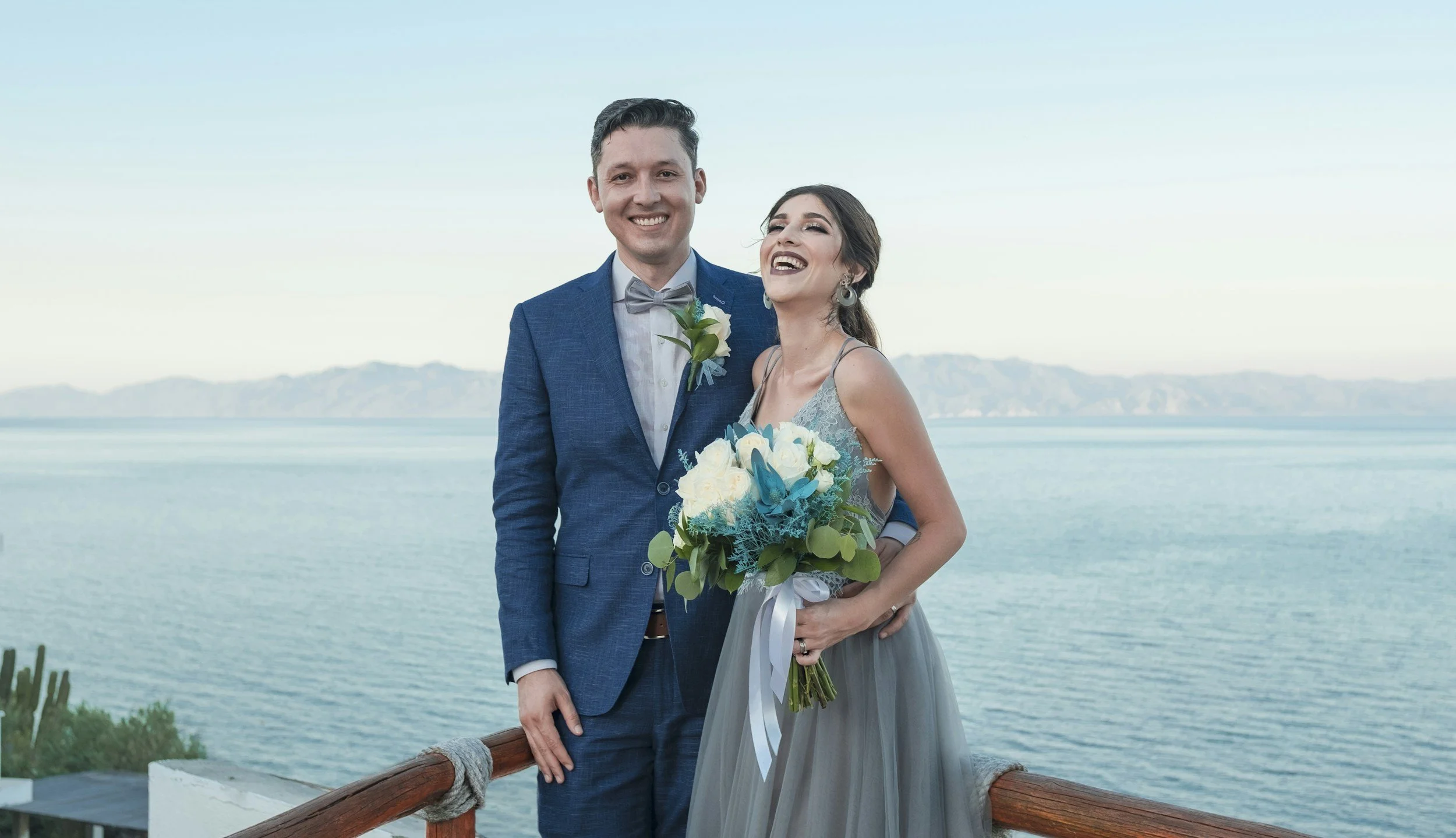 Smiling bride and groom standing on a balcony overlooking the sea, holding a blue and white wedding bouquet.