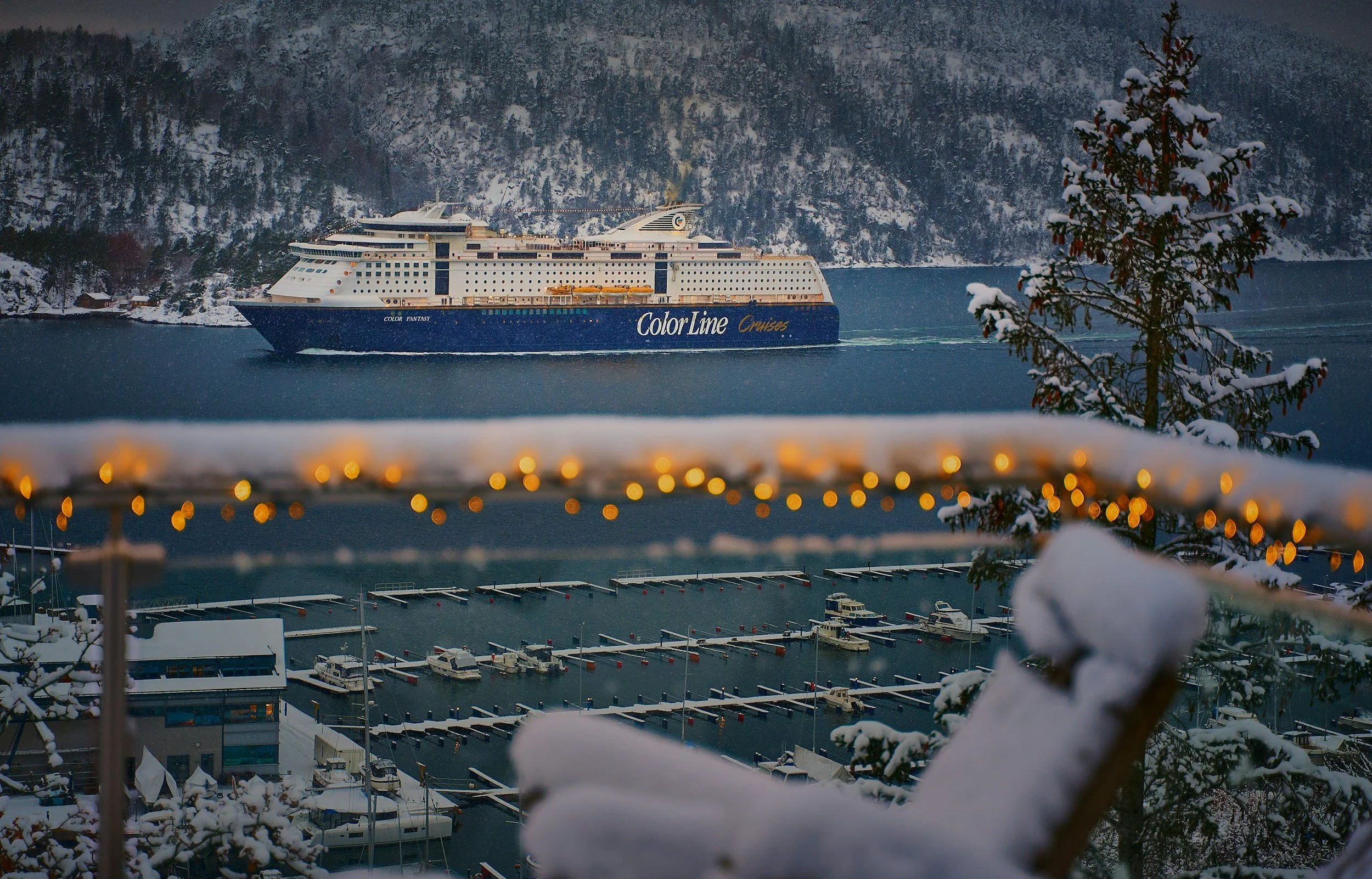 Snowy fjord scene with a Color Line cruise ship sailing past a marina, holiday lights glowing in the foreground.