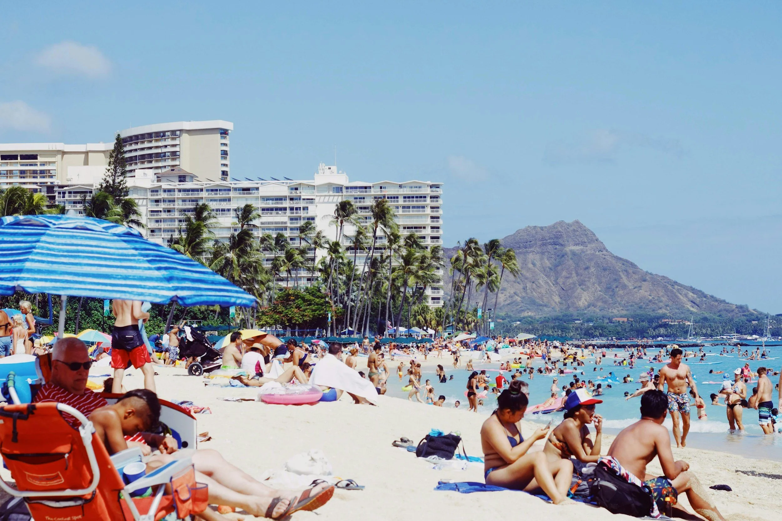 Sun-soaked crowd relaxing on a busy Waikiki Beach with high-rise hotels, palm trees, and Diamond Head crater in the background.