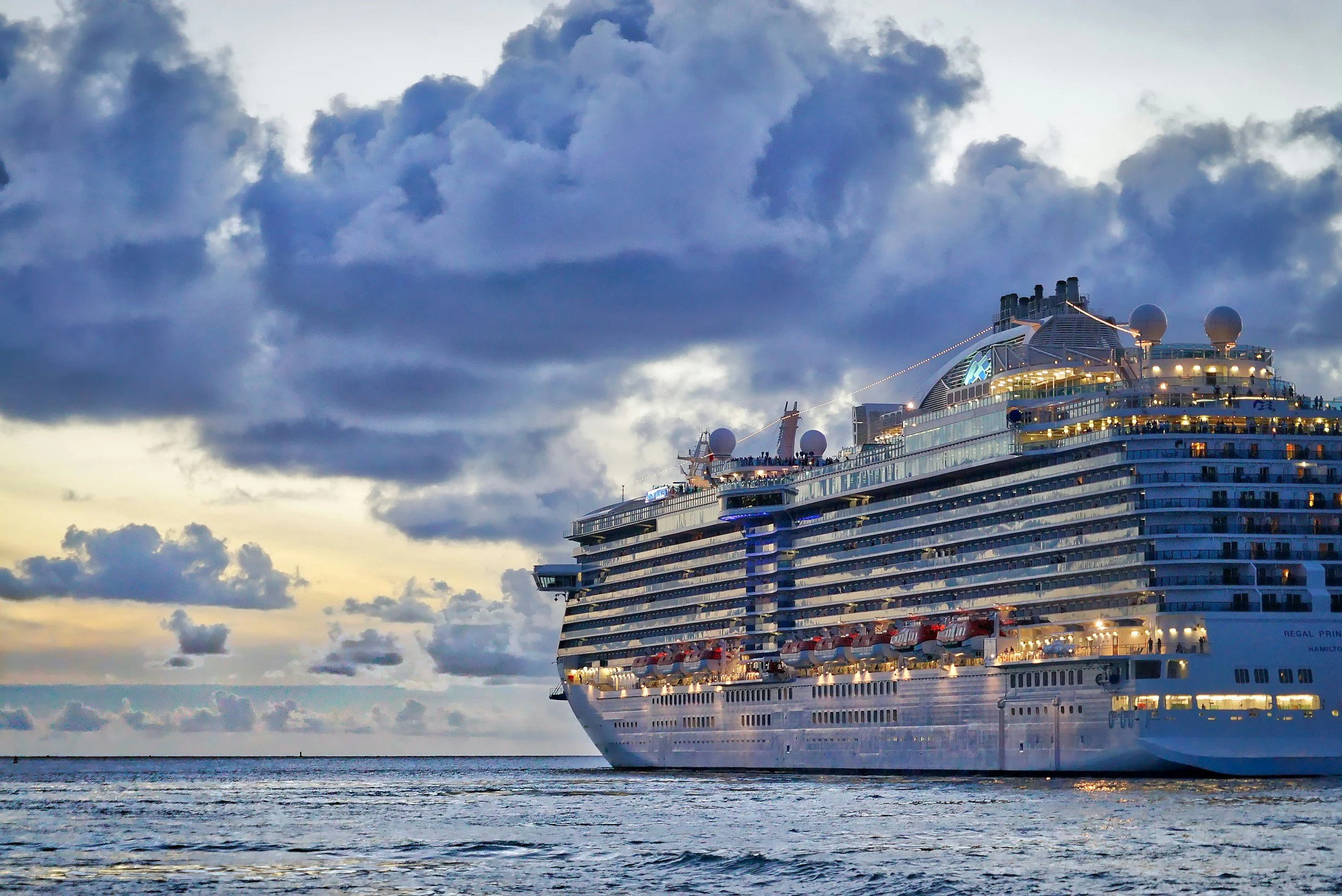 Cruise ship sailing at sunset with dramatic clouds over the ocean and deck lights glowing.