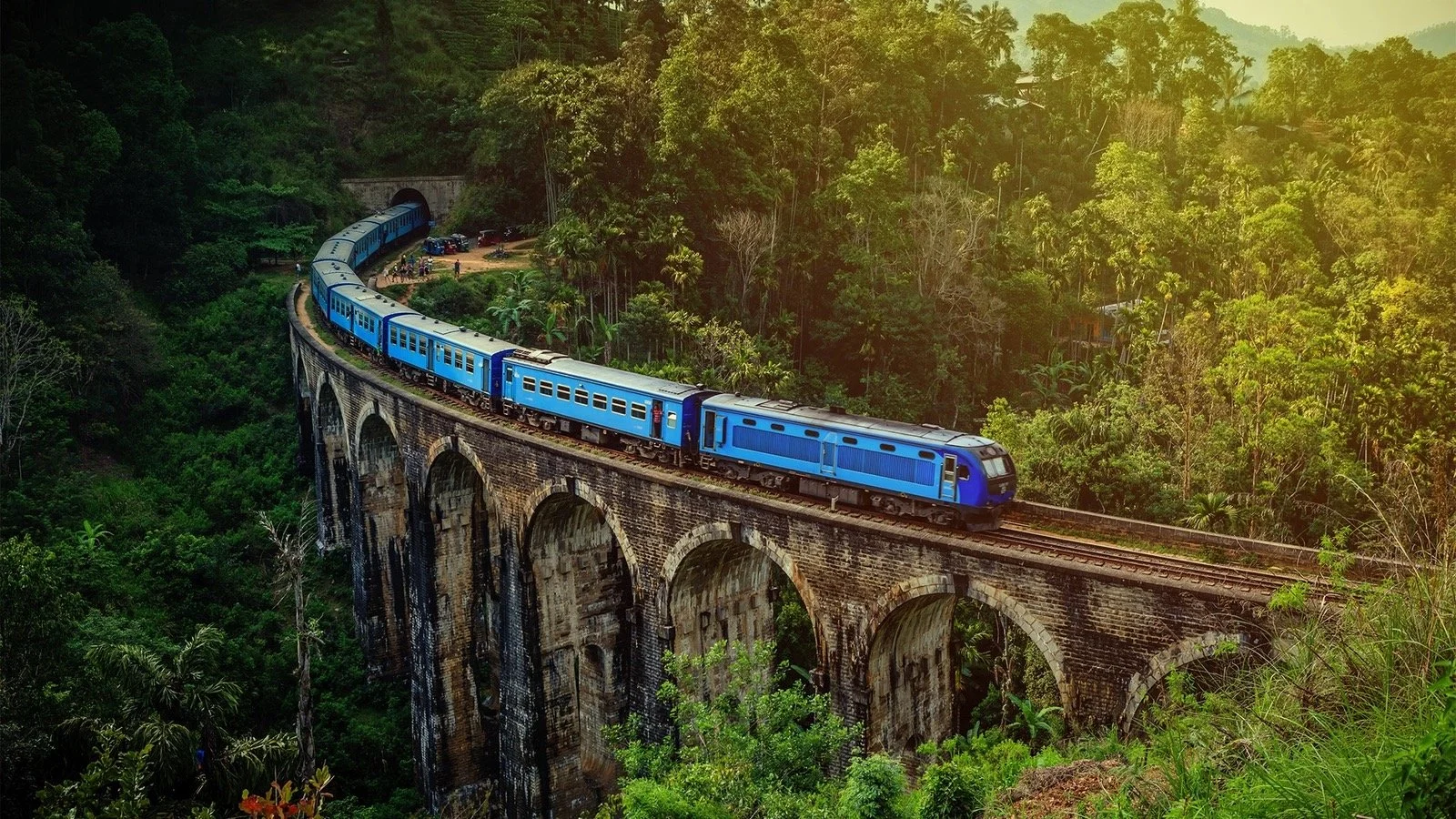 A blue train traveling across a stone arch railway trestle bridge through a lush, green forest.