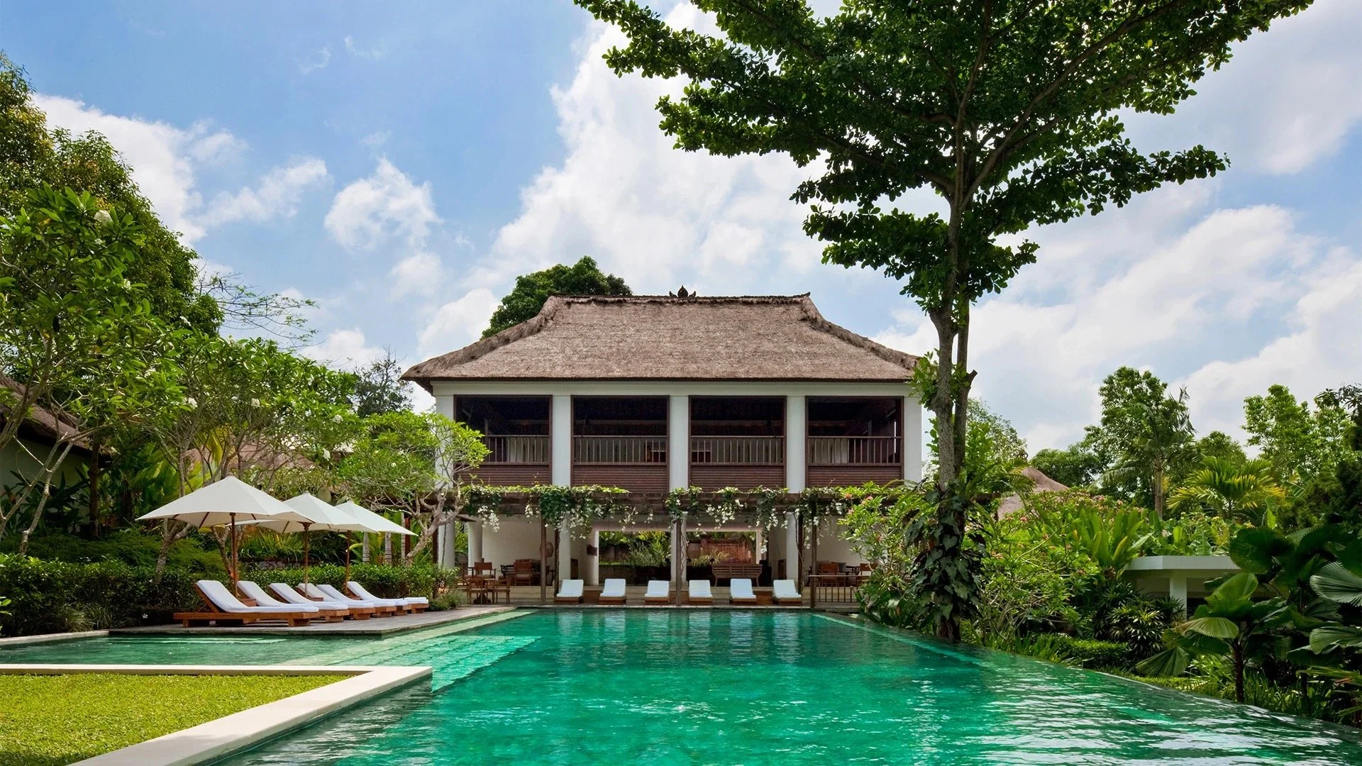 Luxury resort with a large swimming pool, poolside lounge chairs with umbrellas, surrounded by lush green trees and tropical plants, two-story building with a thatched roof and balcony, under a partly cloudy sky.