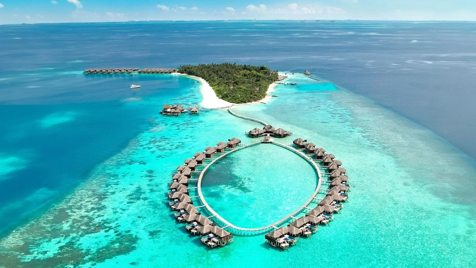 An aerial view of an island with overwater bungalows arranged in a loop and a heart shape, surrounded by turquoise ocean waters and a dense forest on the island.
