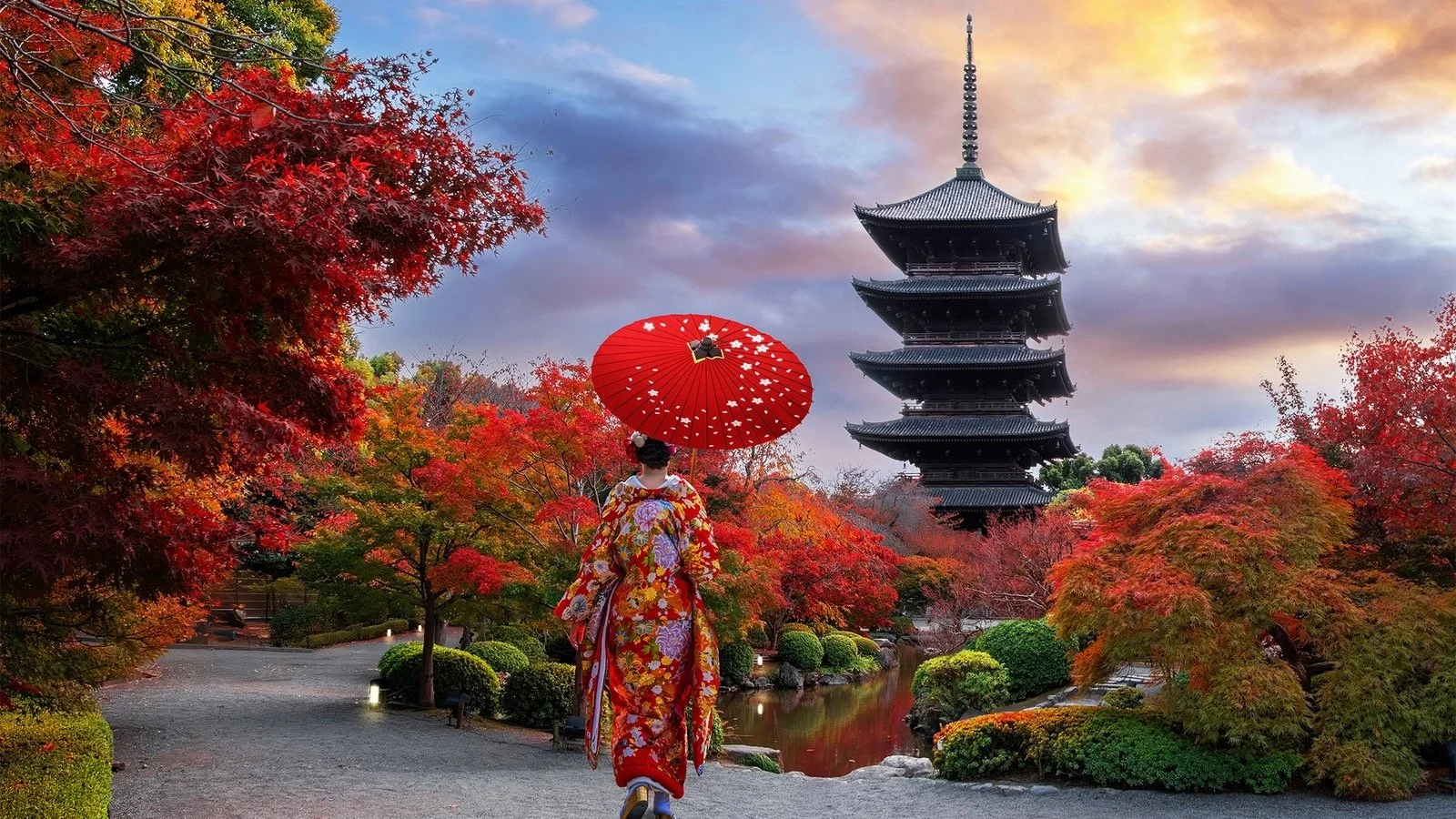 A person dressed in traditional Japanese kimono holding a red umbrella walking through a garden with colorful autumn trees and a tall pagoda in the background at sunset.