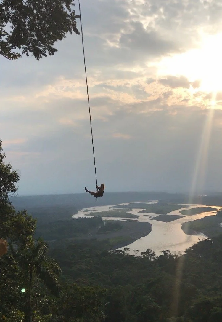 Person swinging on a rope over a scenic river and forest landscape at sunset.