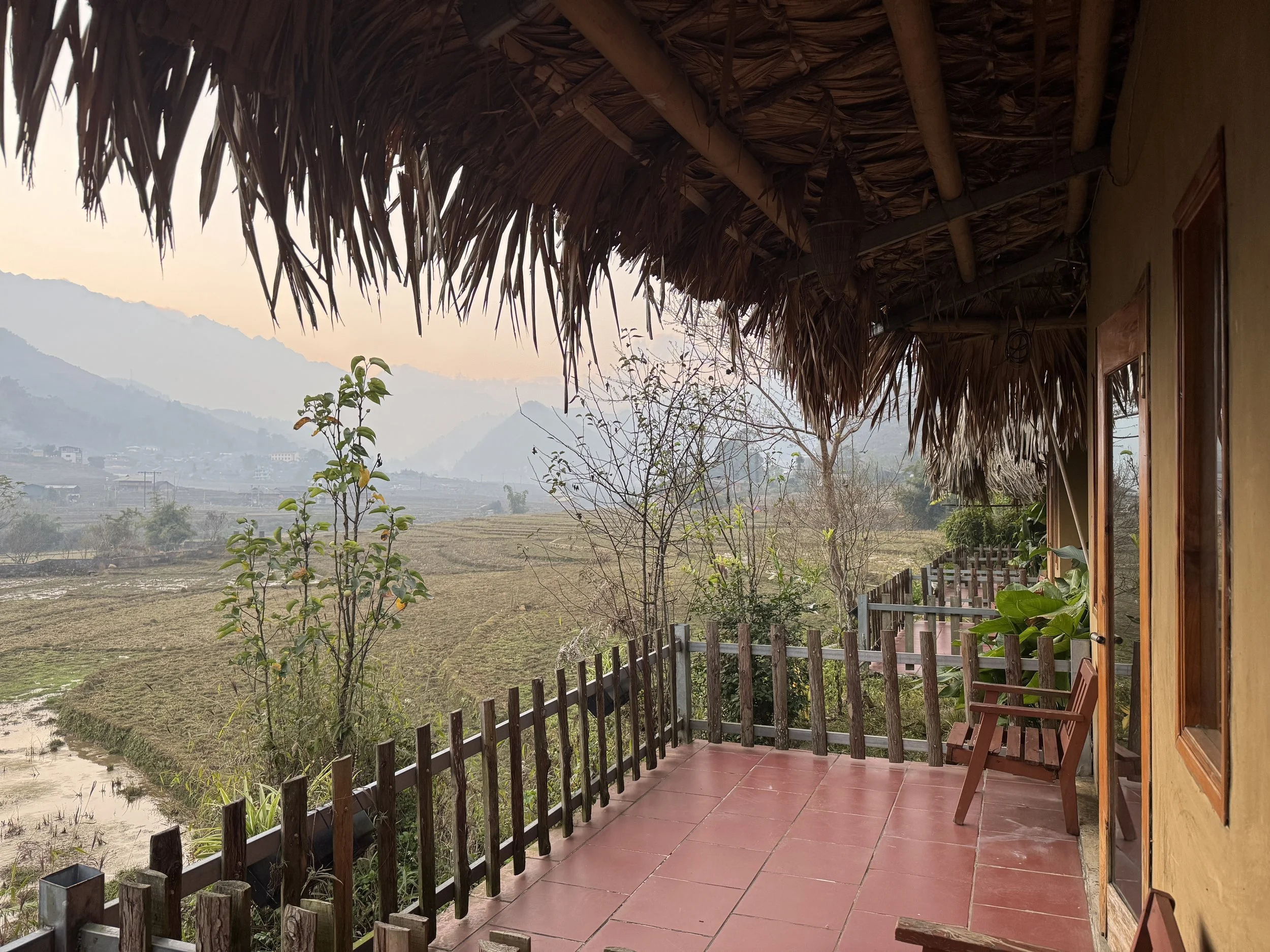 Terrace with thatched roof overlooking scenic rural landscape with fields and mountains in the distance.