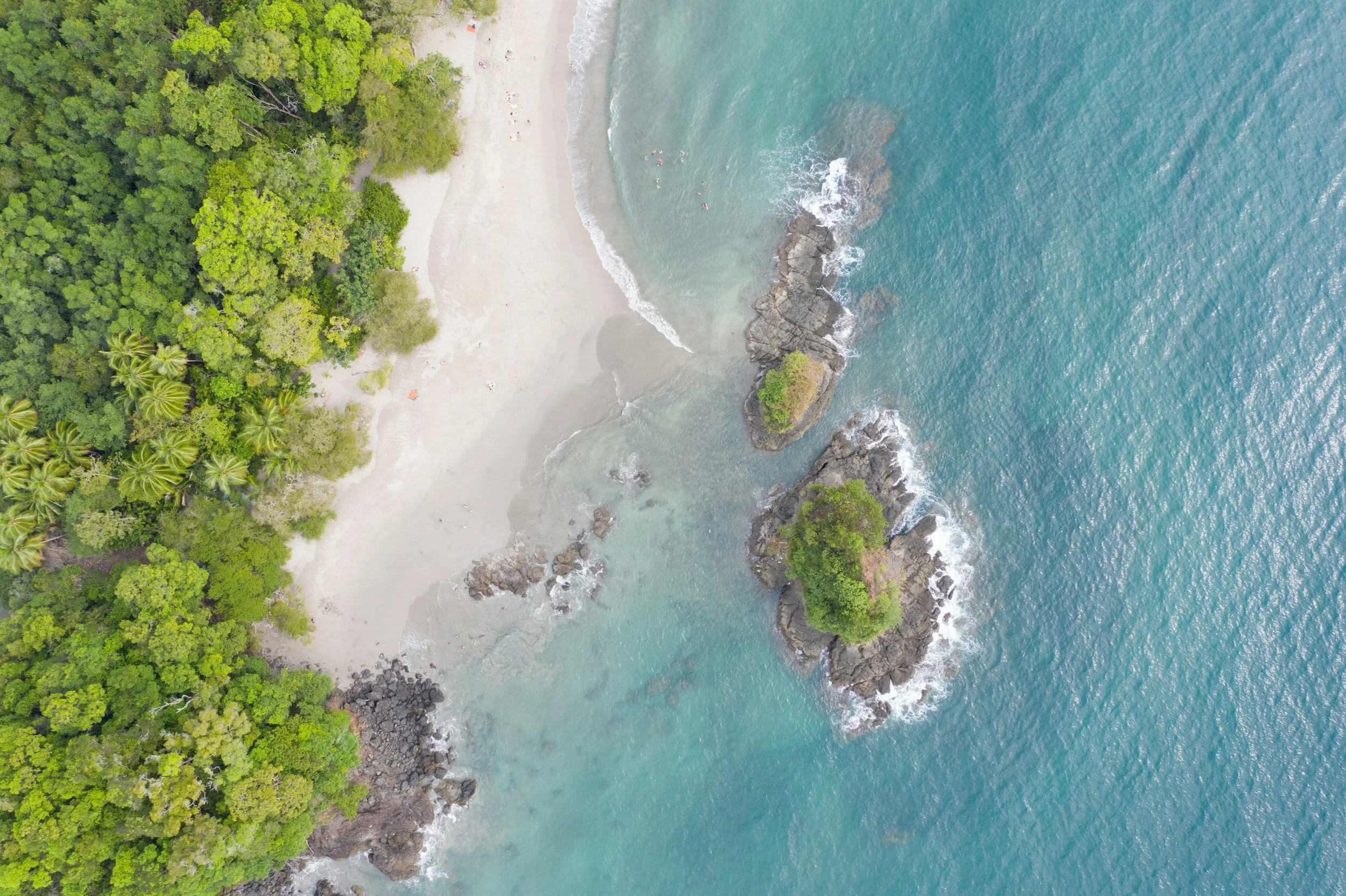 Aerial view of a beach with turquoise water, lush green forest, sandy shores, and rocky outcroppings.