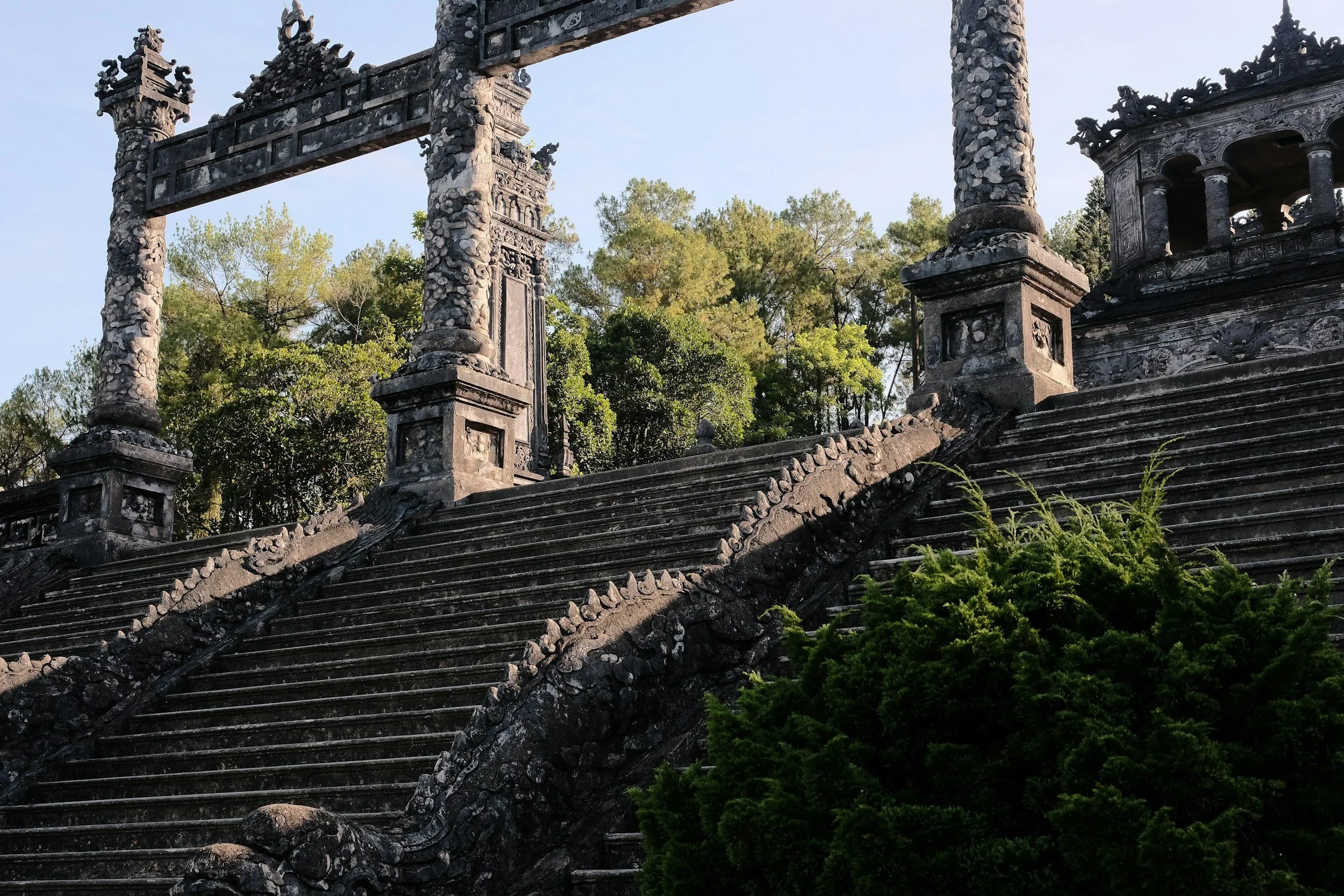 Stone staircase leading to an ornate, historic arch and structure with columns, surrounded by dense greenery and trees.