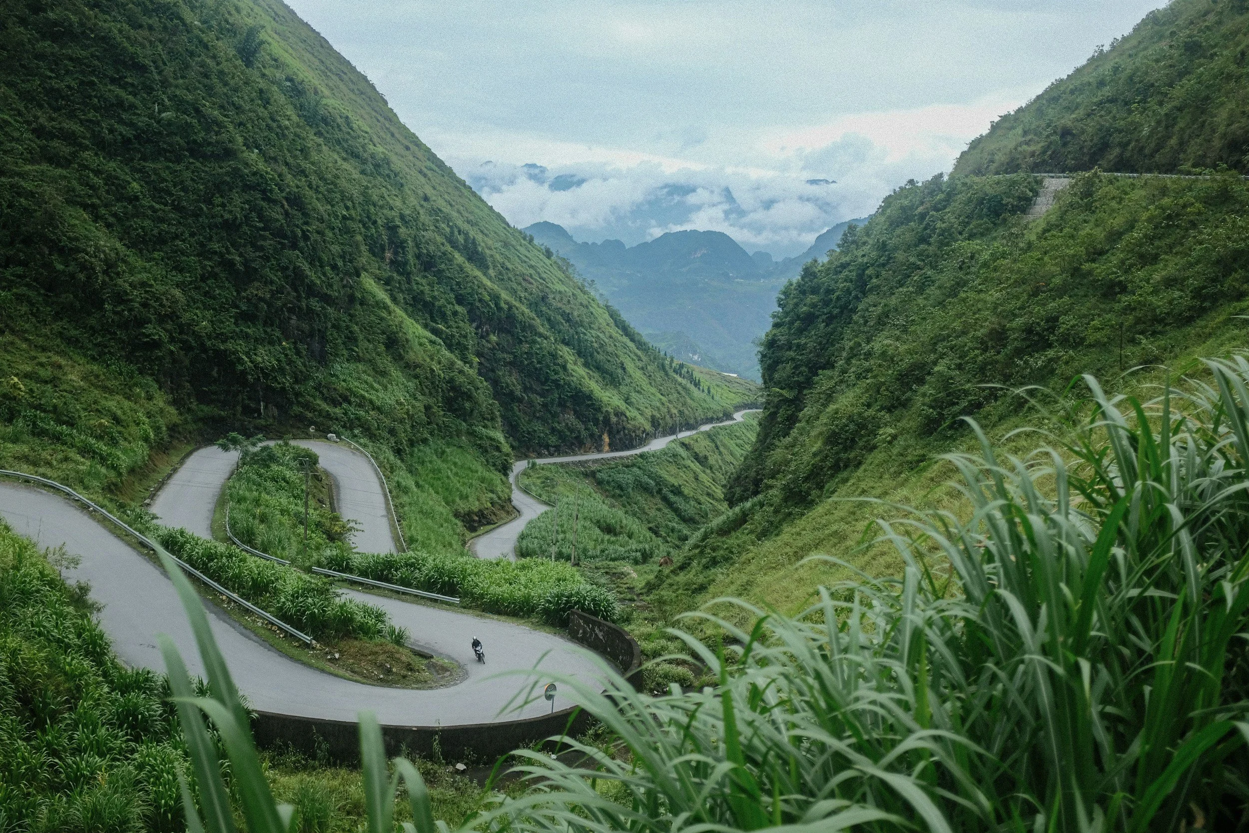 Winding mountain road through lush green hills with a distant view of misty mountains.