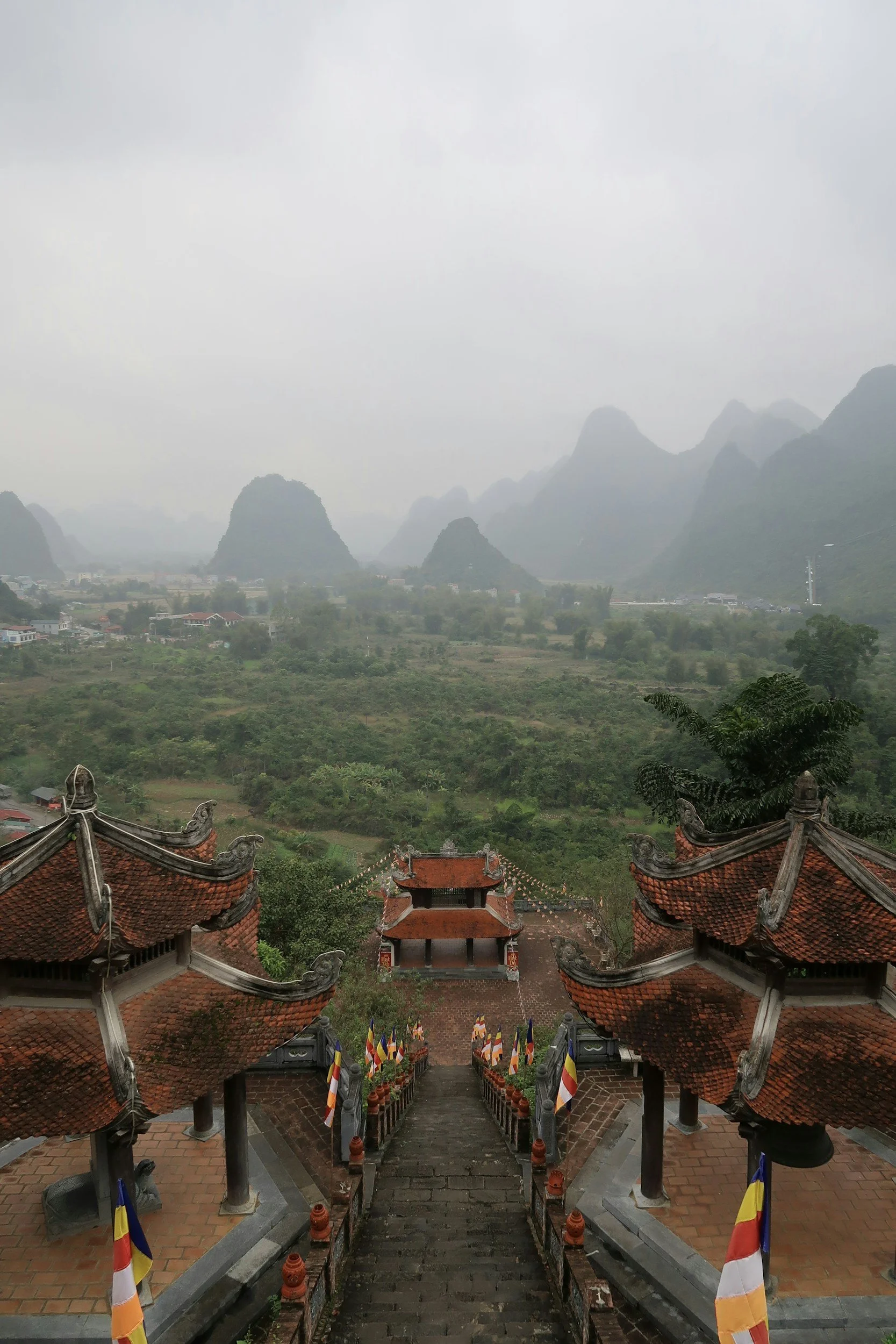Stairway with red-roofed temples, surrounded by lush greenery and misty karst mountains in the background.