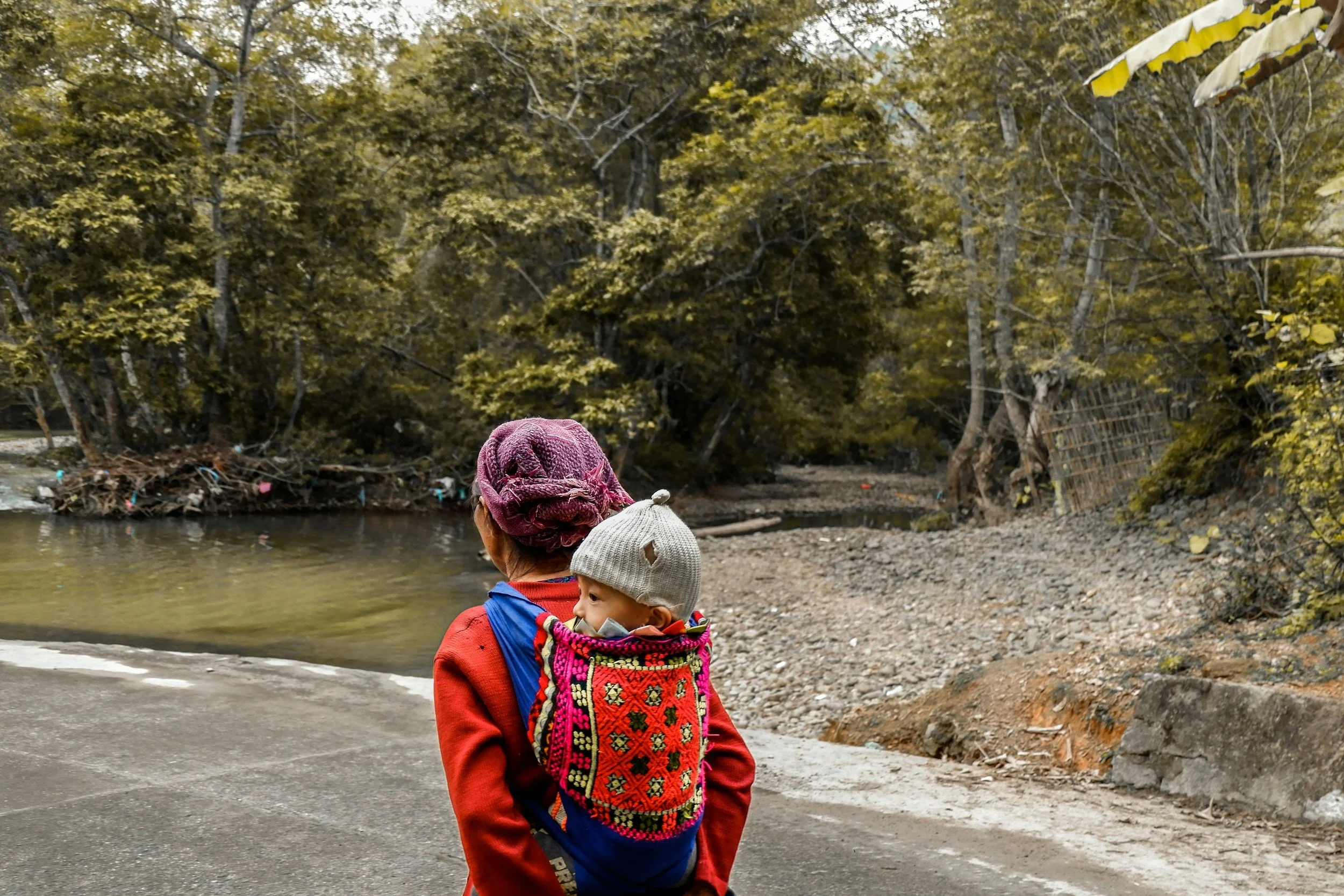 Person carrying a baby in a colorful woven carrier, walking near a river and surrounded by trees.