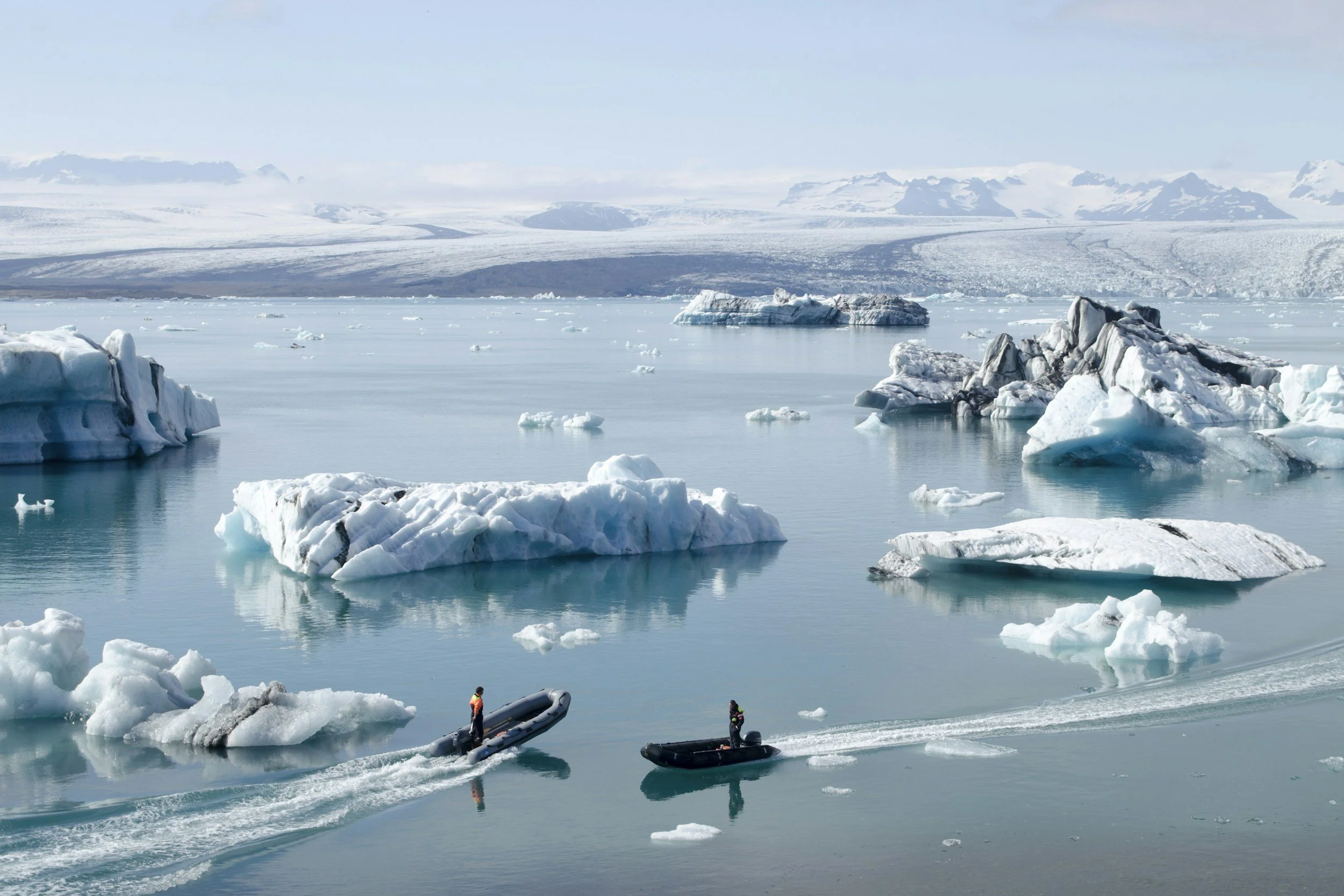 Icebergs floating in a glacier lagoon with two boats and people wearing cold-weather gear. Snow-covered mountains are visible in the background under a clear sky.