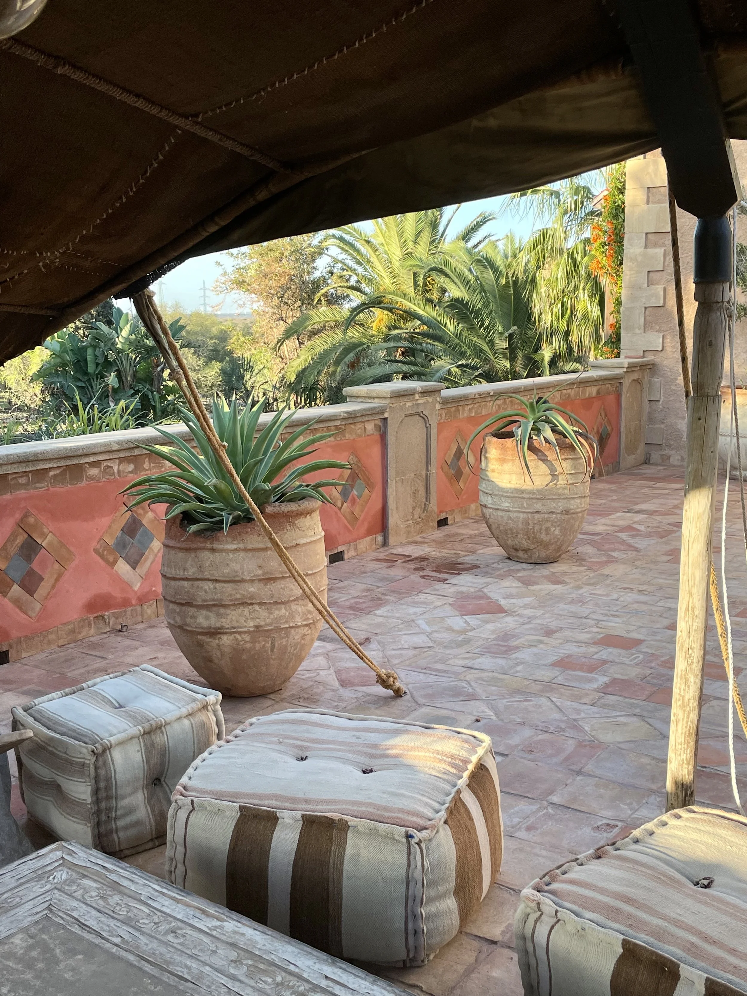 Outdoor terrace with striped cushions, large clay pots with plants, stone tiles, and tropical trees in the background.
