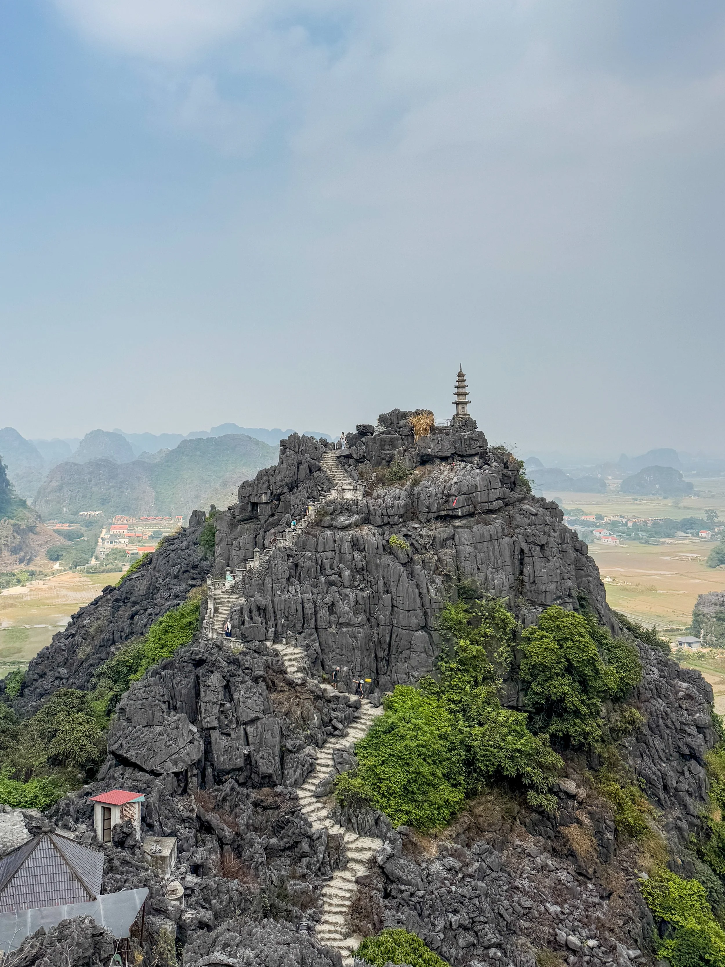 Rocky mountain with stone steps leading to a small pagoda at the summit, surrounded by greenery and distant hills under a cloudy sky.