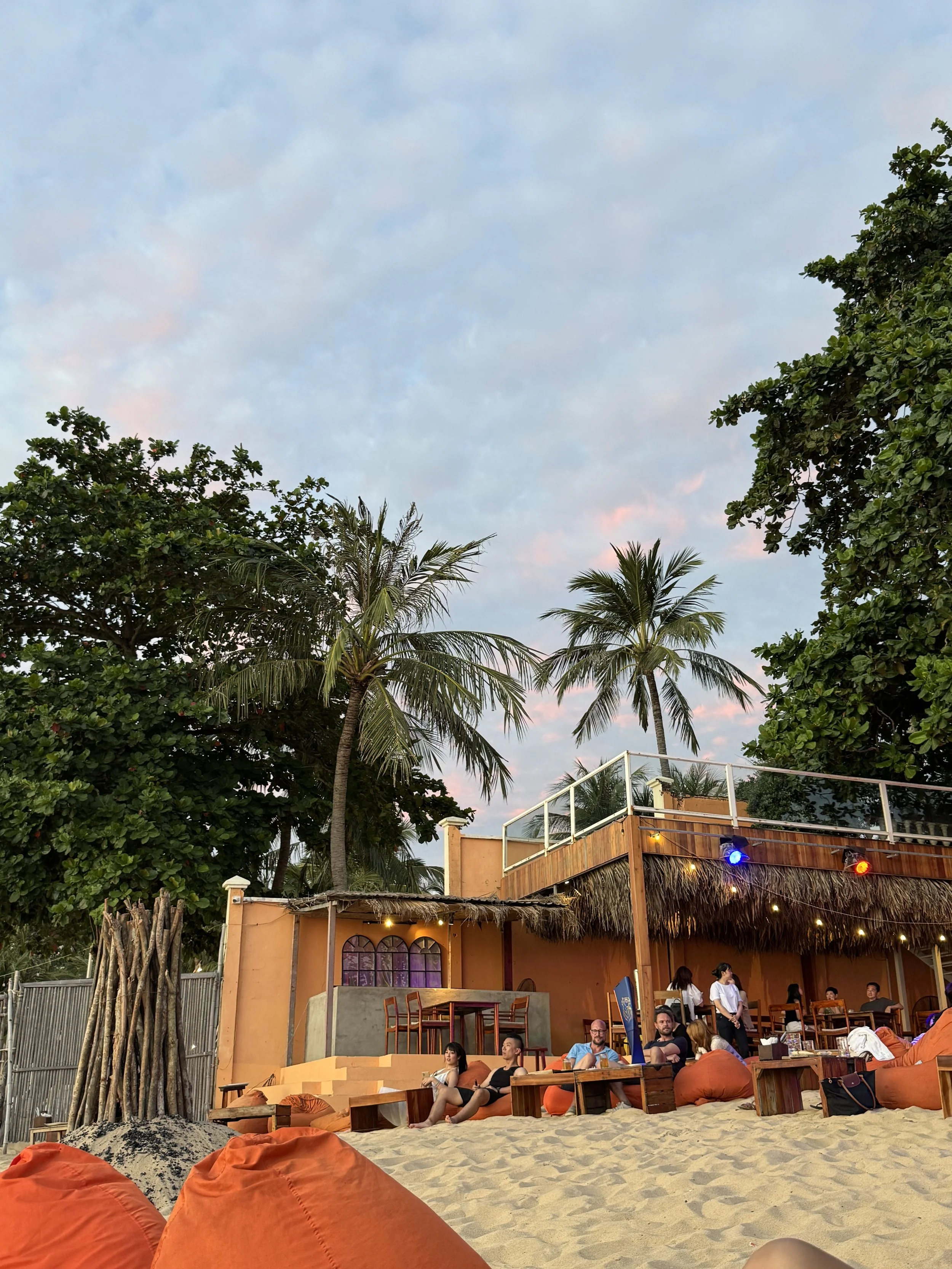 Beachside café with orange bean bags, tables, and people relaxing under palm trees with a thatched roof structure and a vibrant sky.