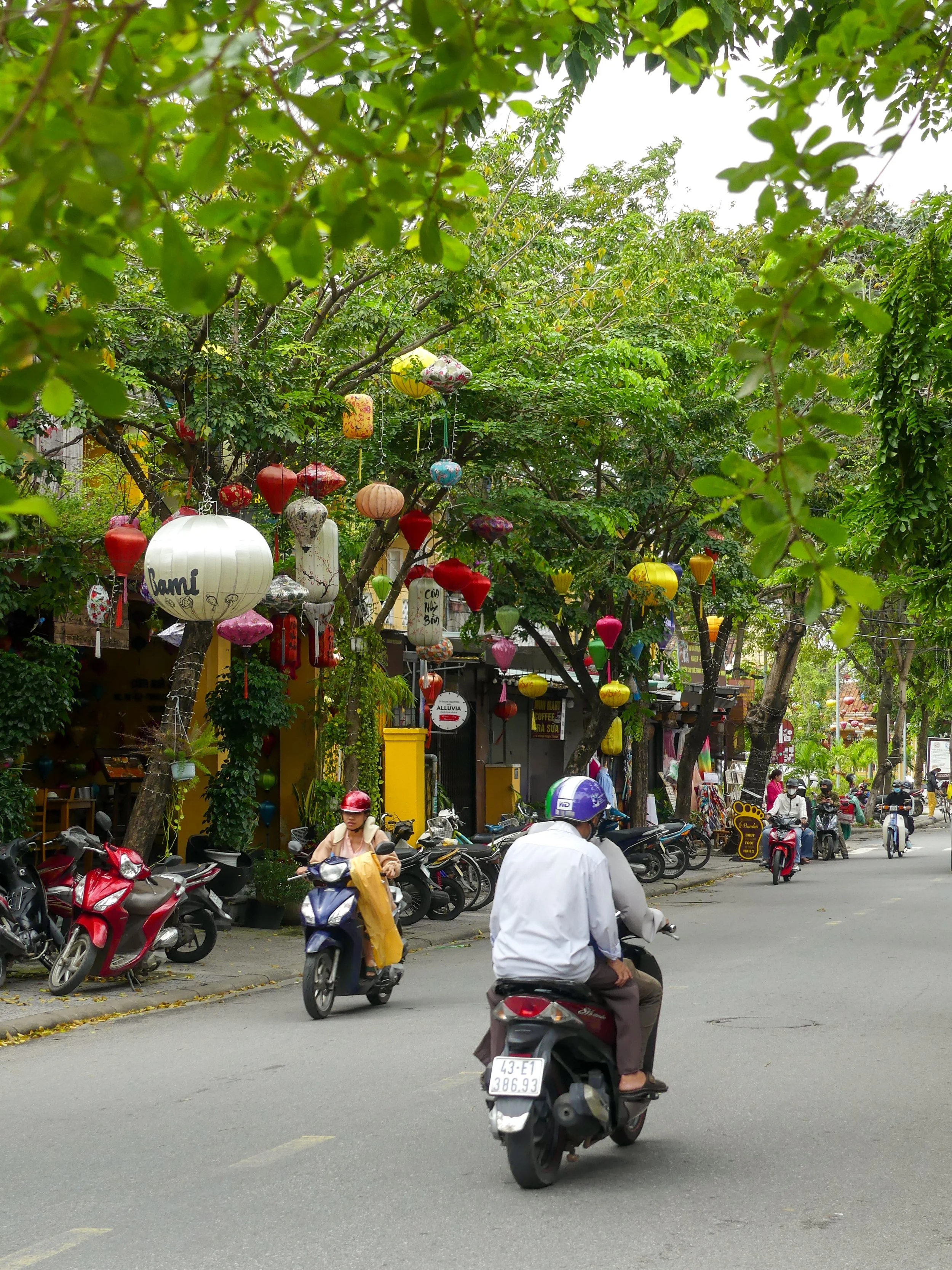 Street scene with people riding motorbikes, colorful hanging lanterns, and parked scooters along the sidewalk, surrounded by lush green trees.