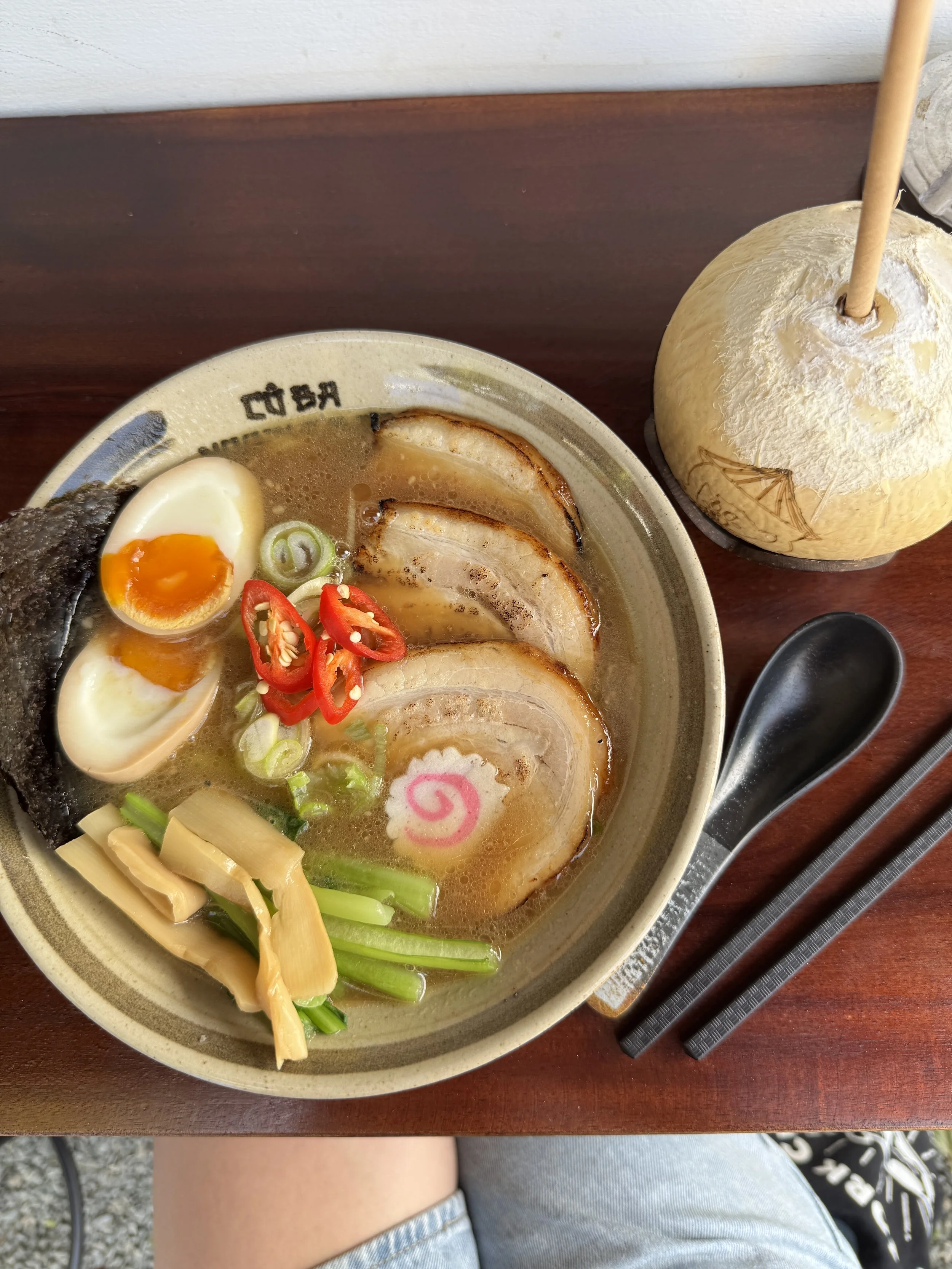Bowl of ramen with pork slices, soft-boiled egg, seaweed, bamboo shoots, green onions, red chili peppers, and narutomaki, accompanied by a coconut drink and black utensils.