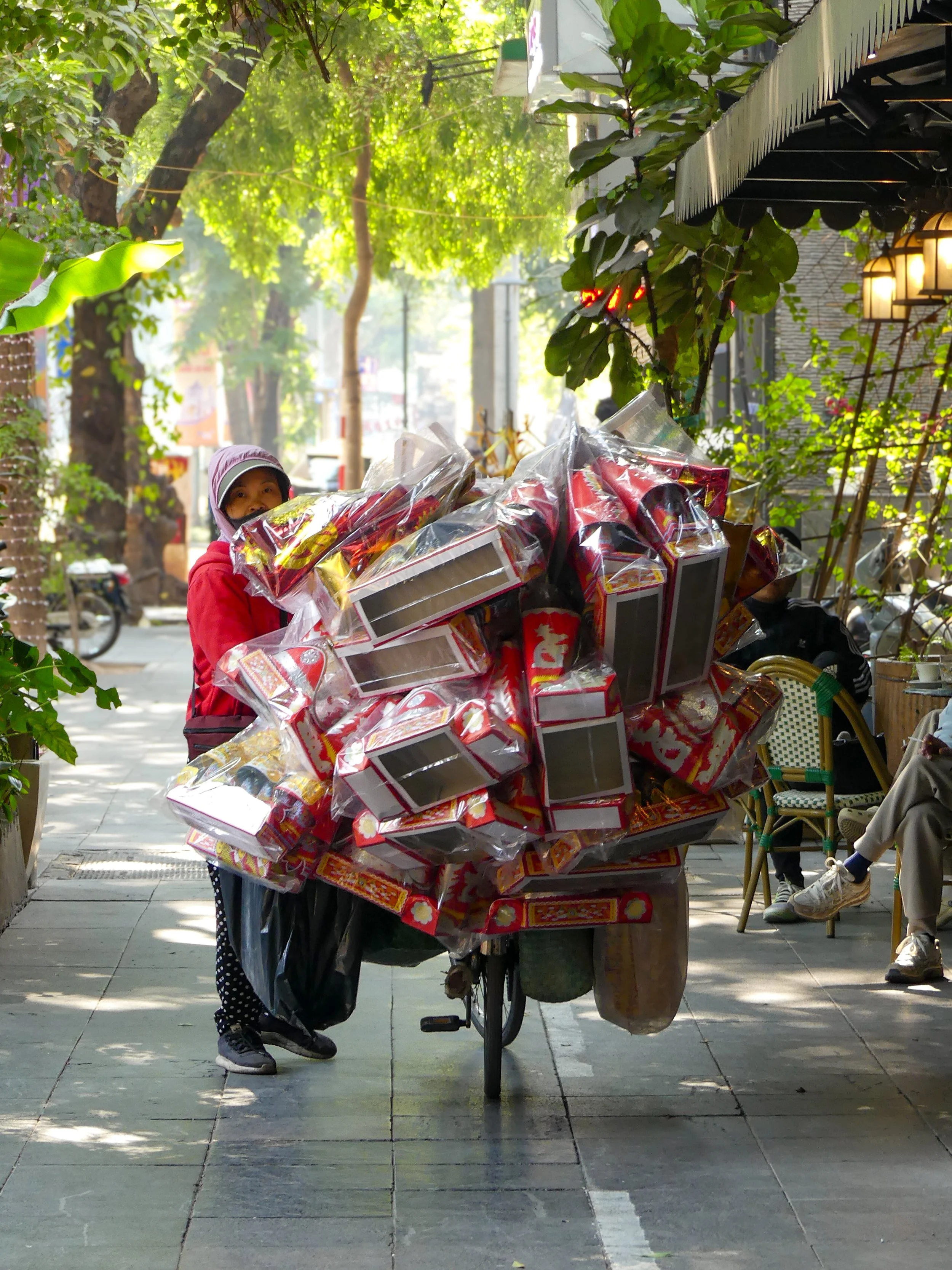 Person on a bicycle loaded with cardboard boxes, wrapped in plastic, in an urban outdoor setting with trees and greenery.
